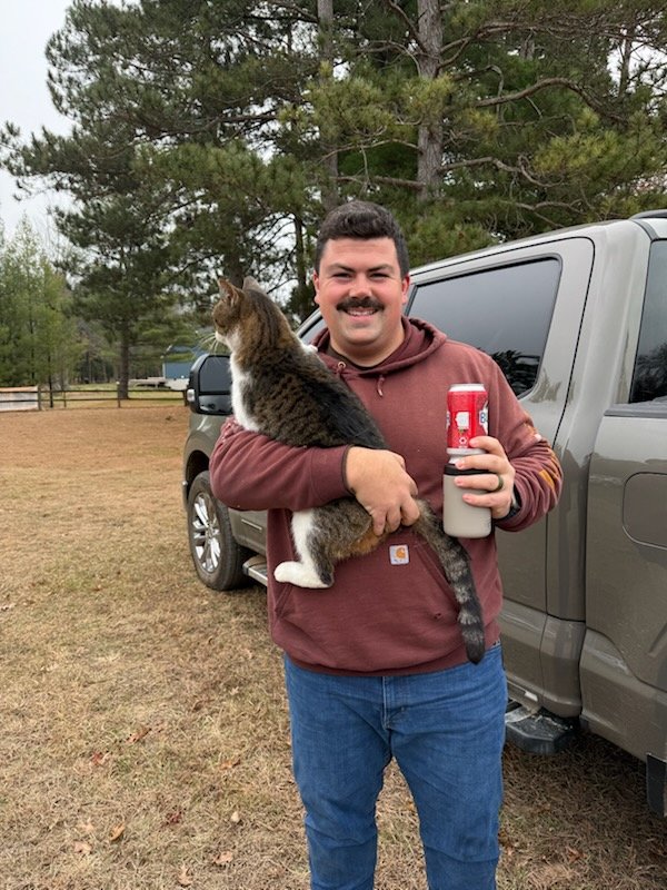 A man with a mustache smiling, holding a large tabby cat in one arm and a soda cup in the other, standing outside near a gray pickup truck in a grassy area with trees.