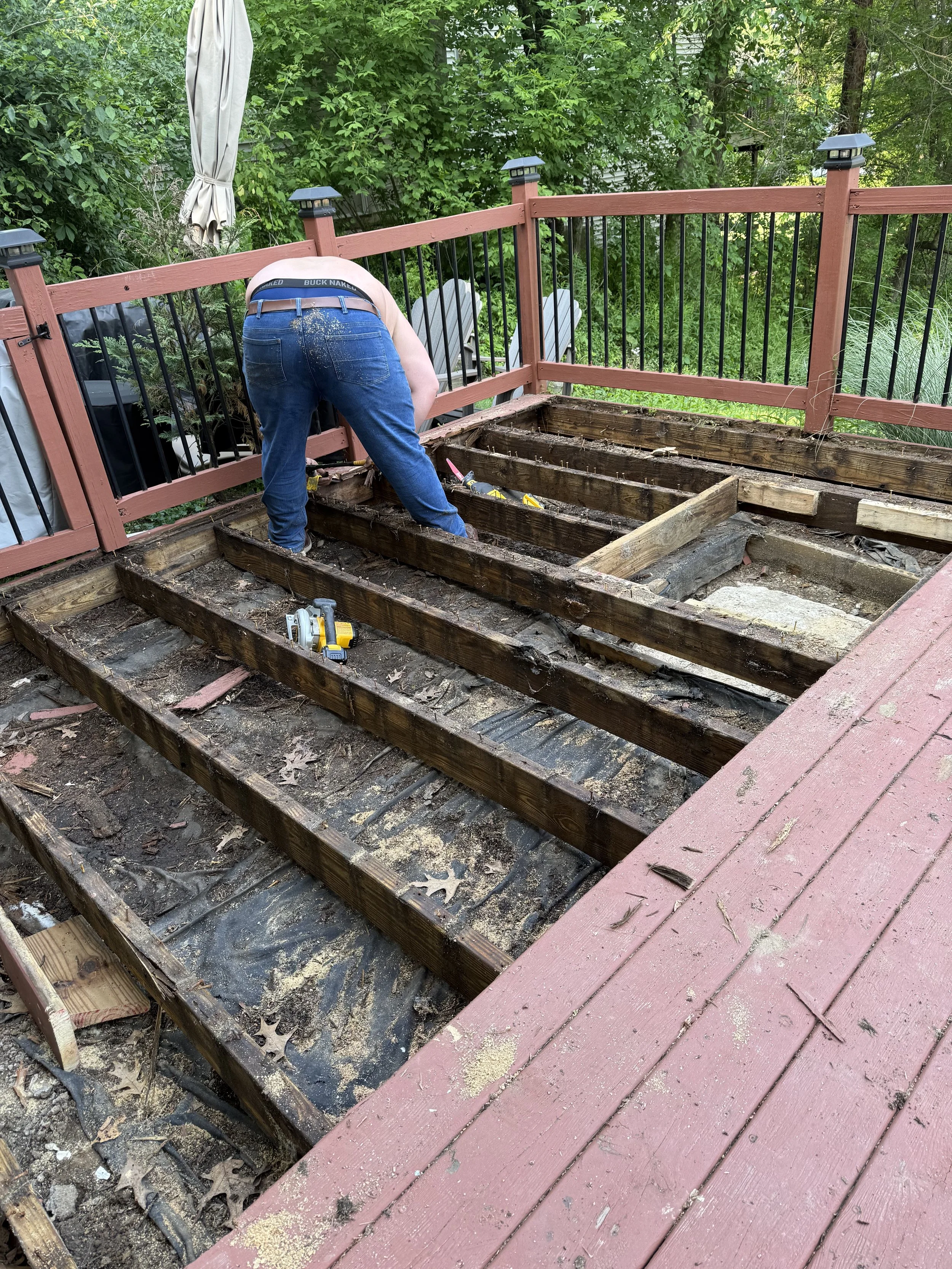 A person working on a deck restoration project, removing old wood and preparing the space for new flooring, with tools and a protective deck railing visible.