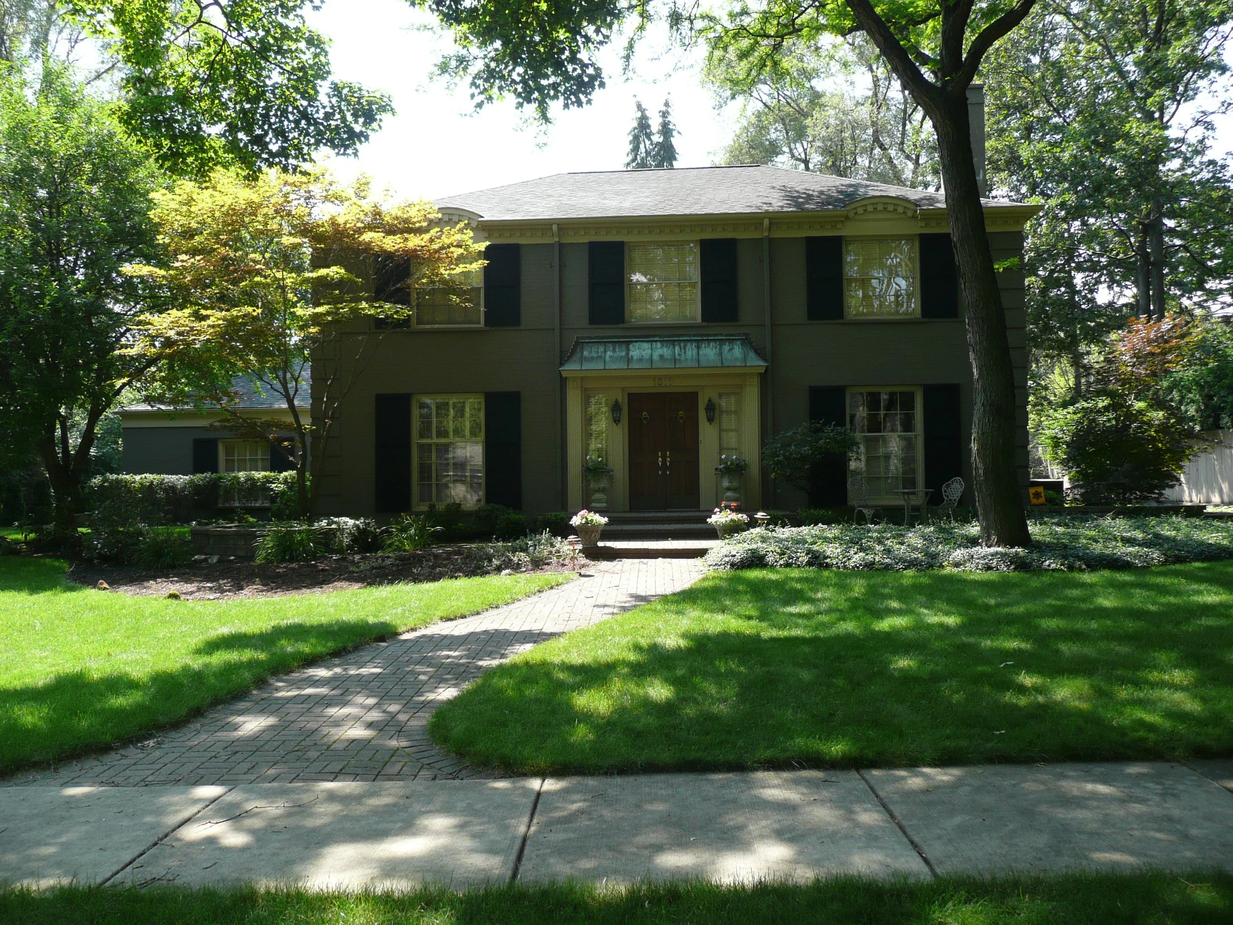 A two-story dark gray house with black shutters, front steps, a small porch, and a green metal awning over the door, surrounded by lush trees and a well-maintained lawn with a brick pathway leading to the front entrance.