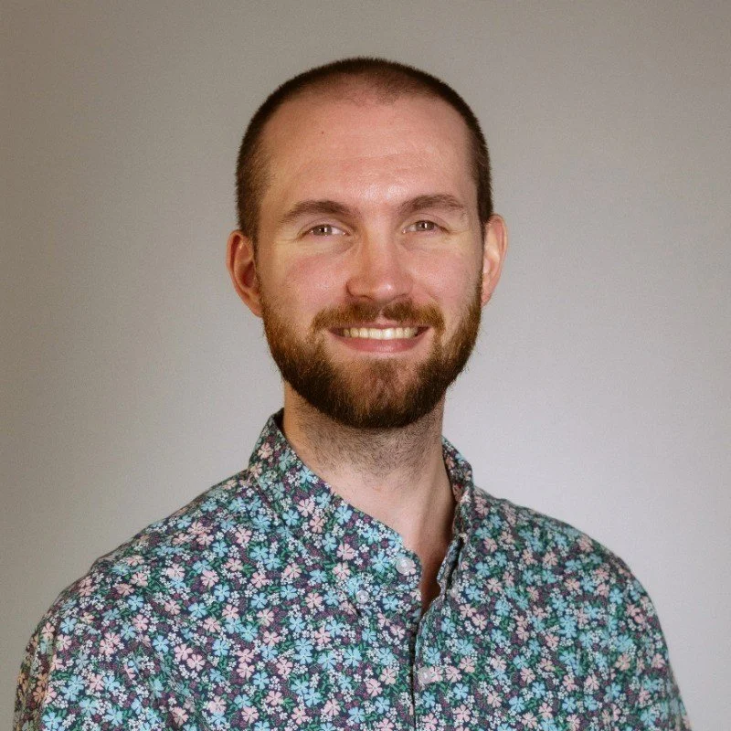 A smiling man with a beard and short hair wearing a floral patterned shirt, standing against a plain background.
