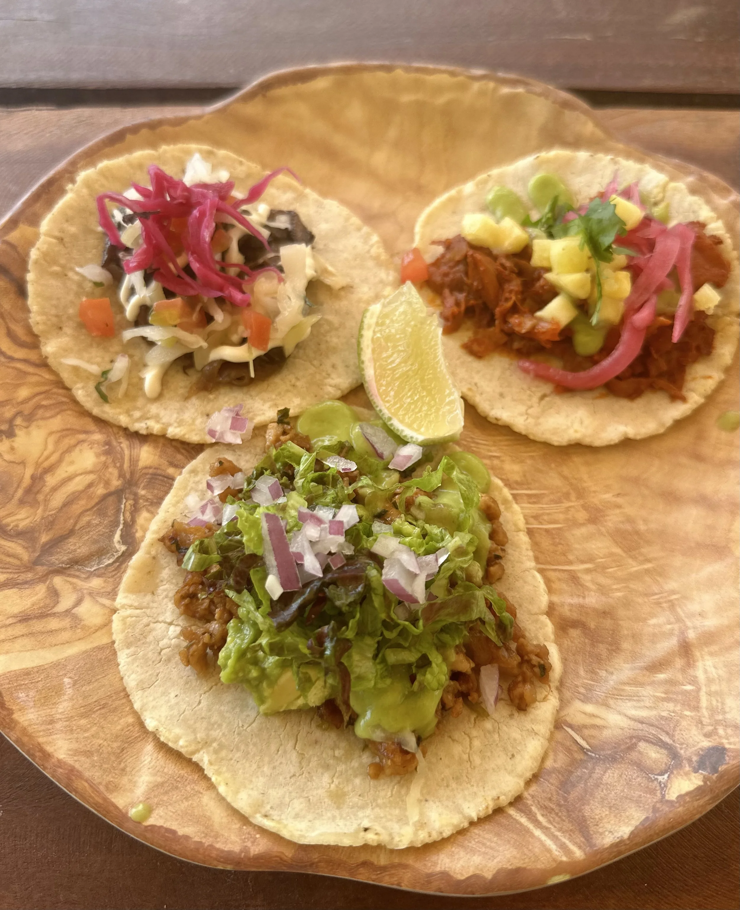 A wooden plate with three small tacos topped with shredded cheese, pickled red onions, chopped vegetables, and a lime wedge.