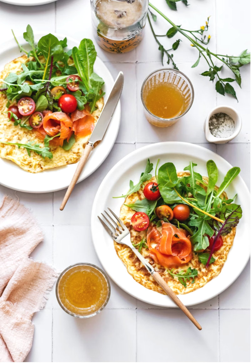 Two plates of omelets topped with cherry tomatoes, mixed greens, and smoked salmon, along with glasses of iced tea and a small jug of honey, on a white table with a pink cloth napkin and green sprigs.