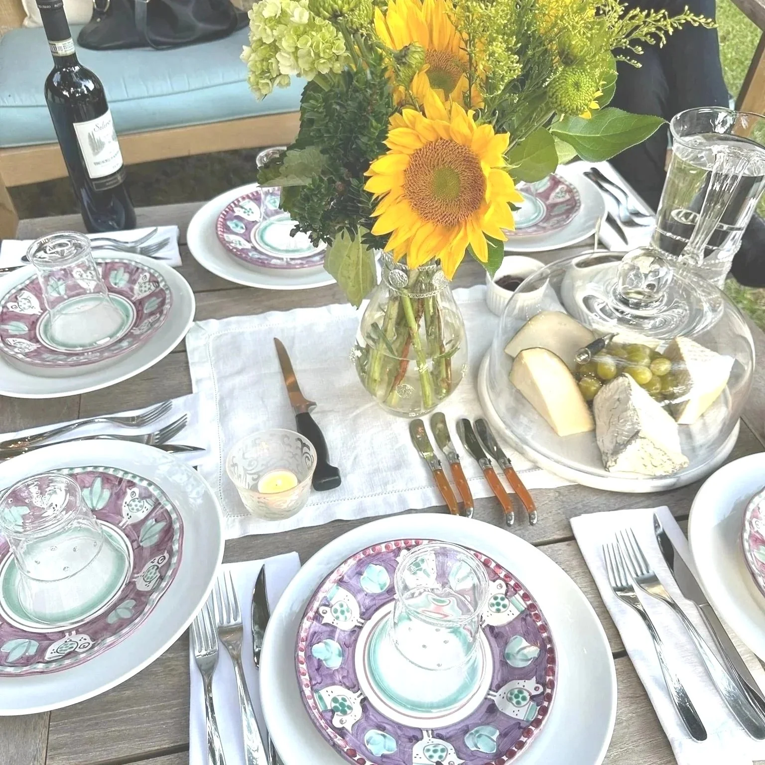 A table setting with plates, forks, knives, and glasses arranged around a wooden table. There is a centerpiece with sunflowers and other flowers in a glass vase, and a platter with cheese, grapes, and crackers. A bottle of wine and a glass of water are also visible.