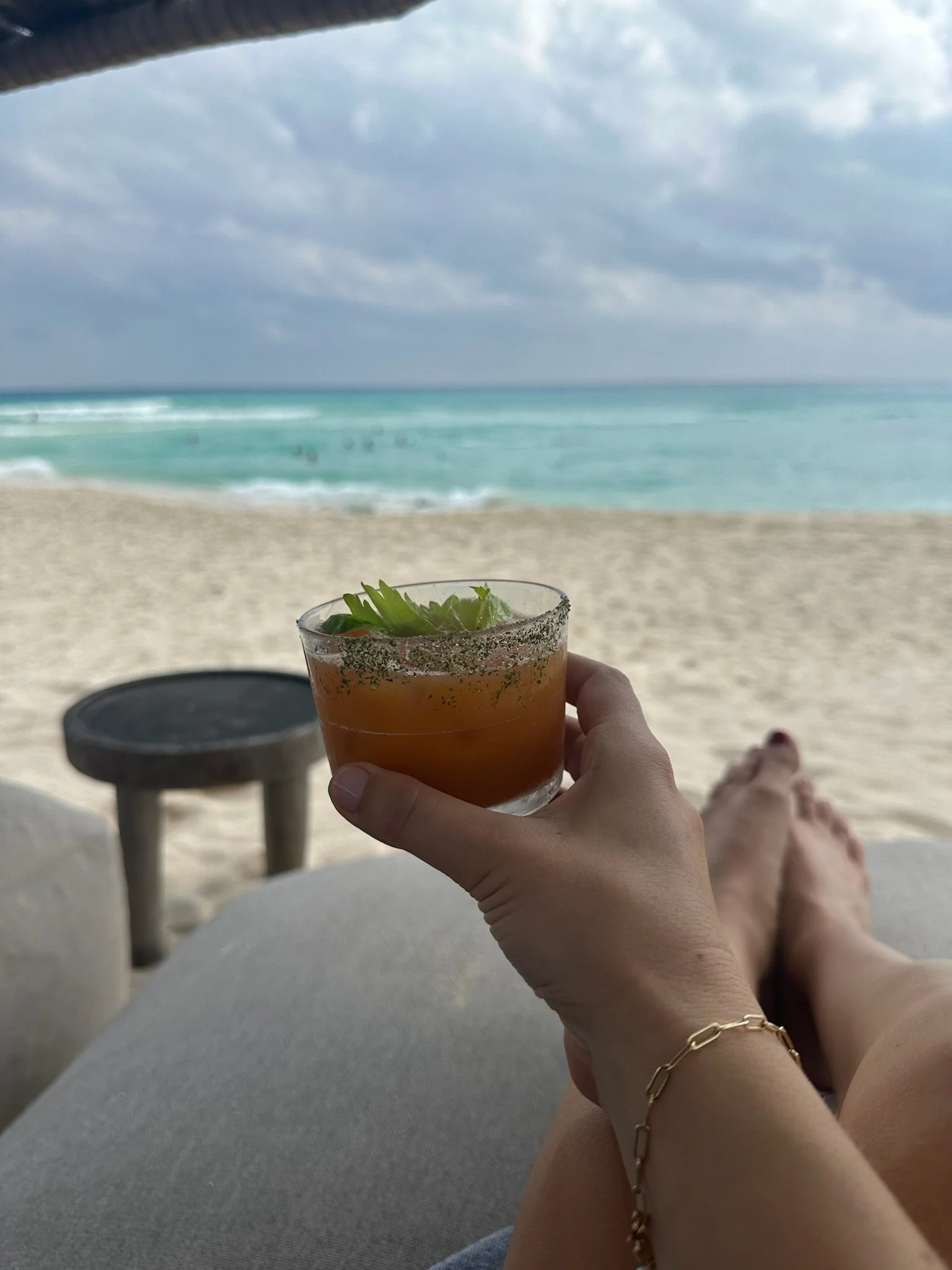 Person relaxing on a beach, holding a cocktail with lime garnishes, with the sea and cloudy sky in the background.