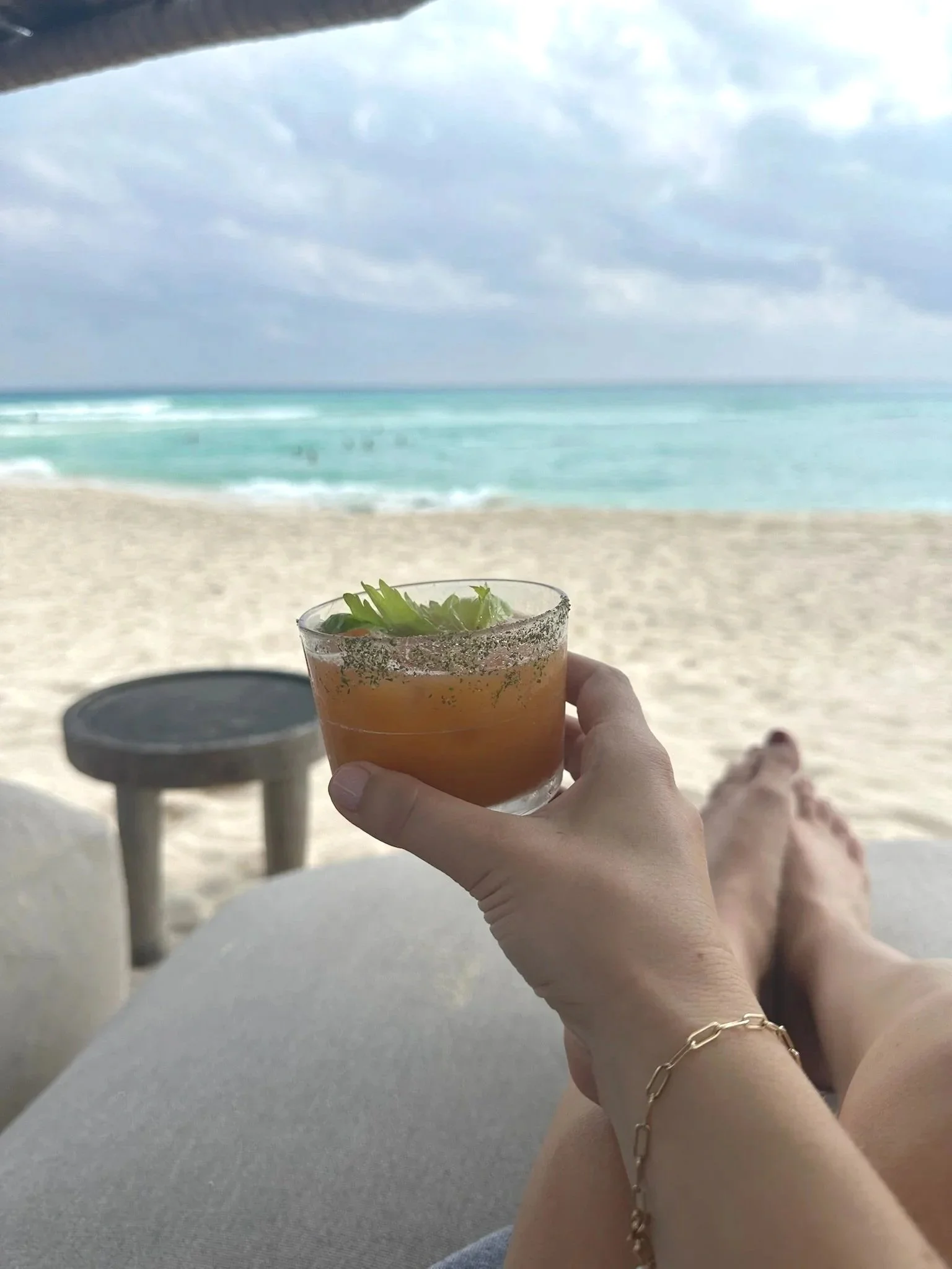 Person relaxing on a lounge chair at the beach, holding a cocktail with a lime garnish, with ocean waves and cloudy sky in the background.