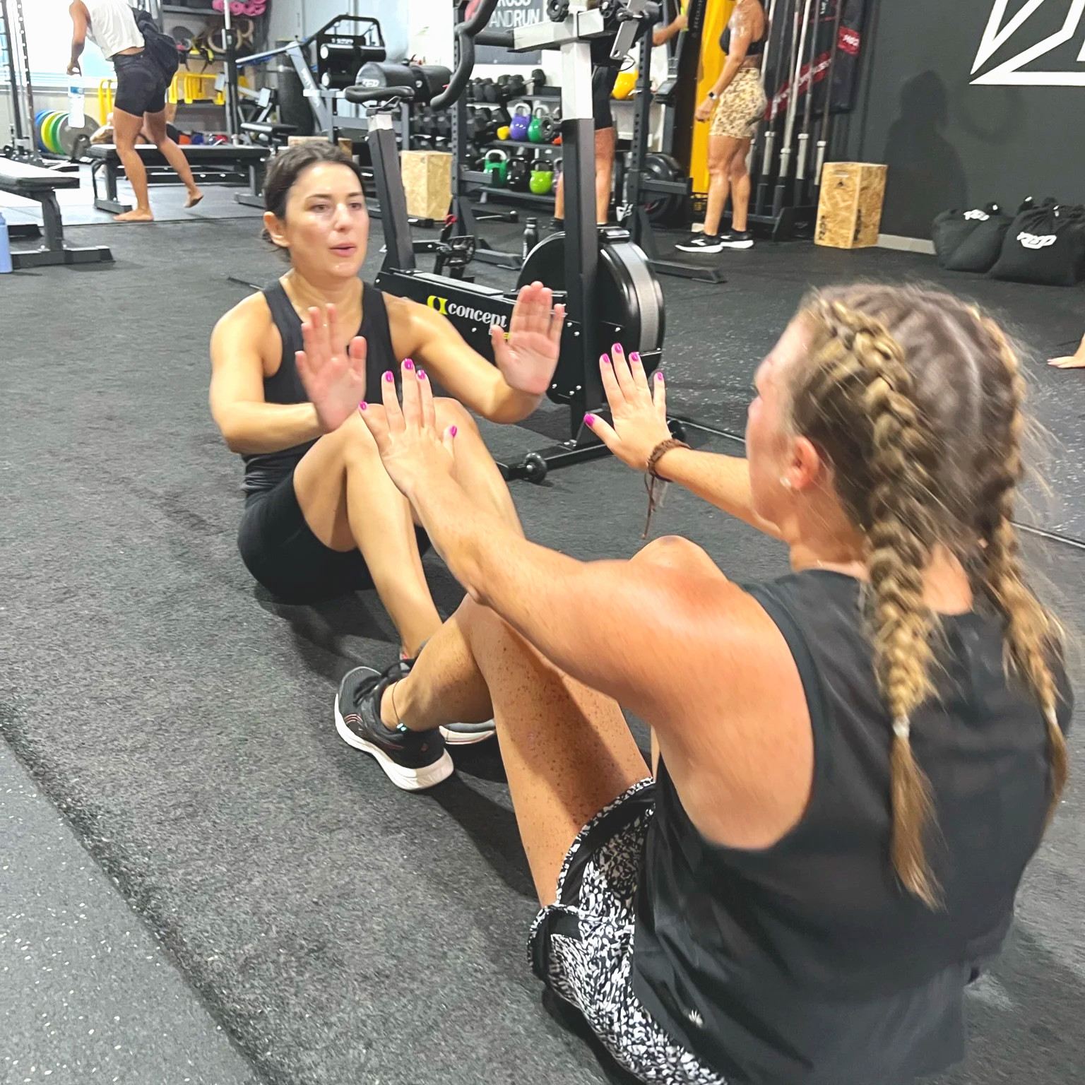 Two women are sitting on the gym floor working facing each other, working out engaging in partner exercises. The gym has exercise equipment in the background, including a stationary bike, weights, and other workout gear.