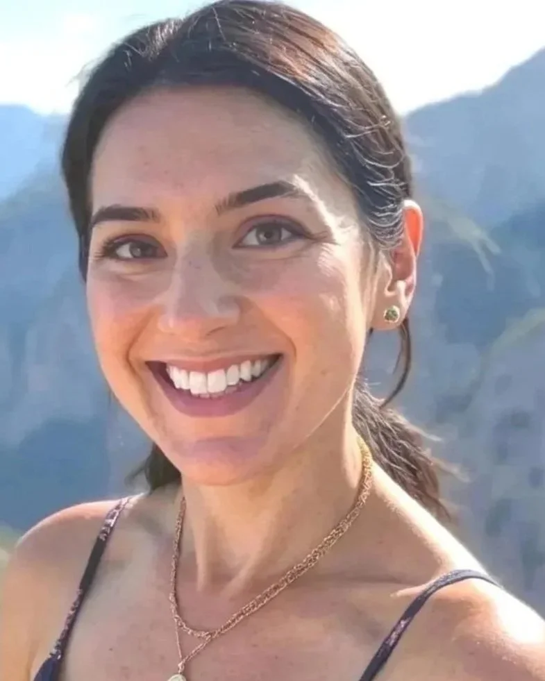 A woman with dark hair tied back, smiling, wearing earrings and necklaces, with mountains and sky in the background.