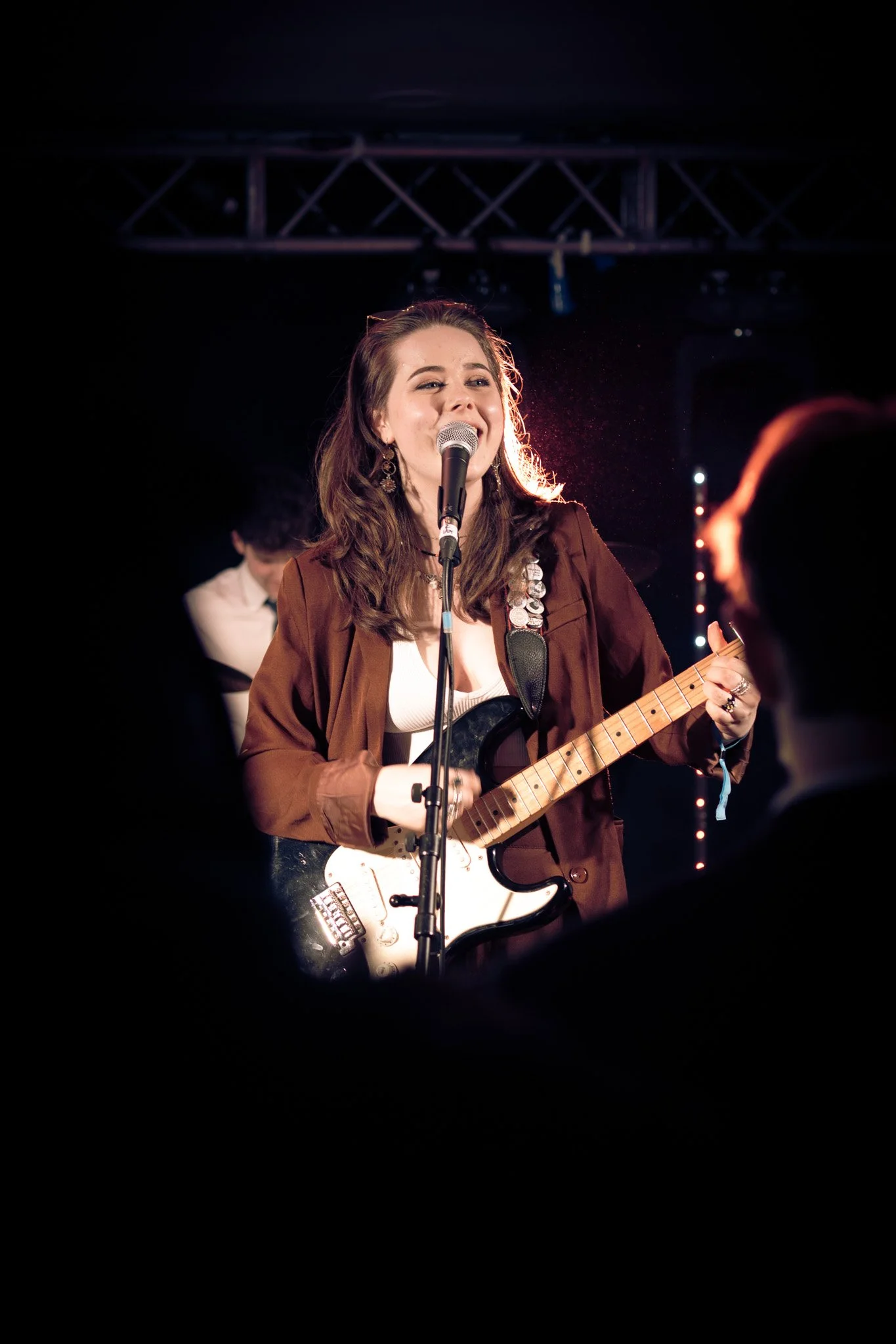 A young woman with brown hair performing on stage, singing into a microphone and playing an electric guitar, with a musician in the background.