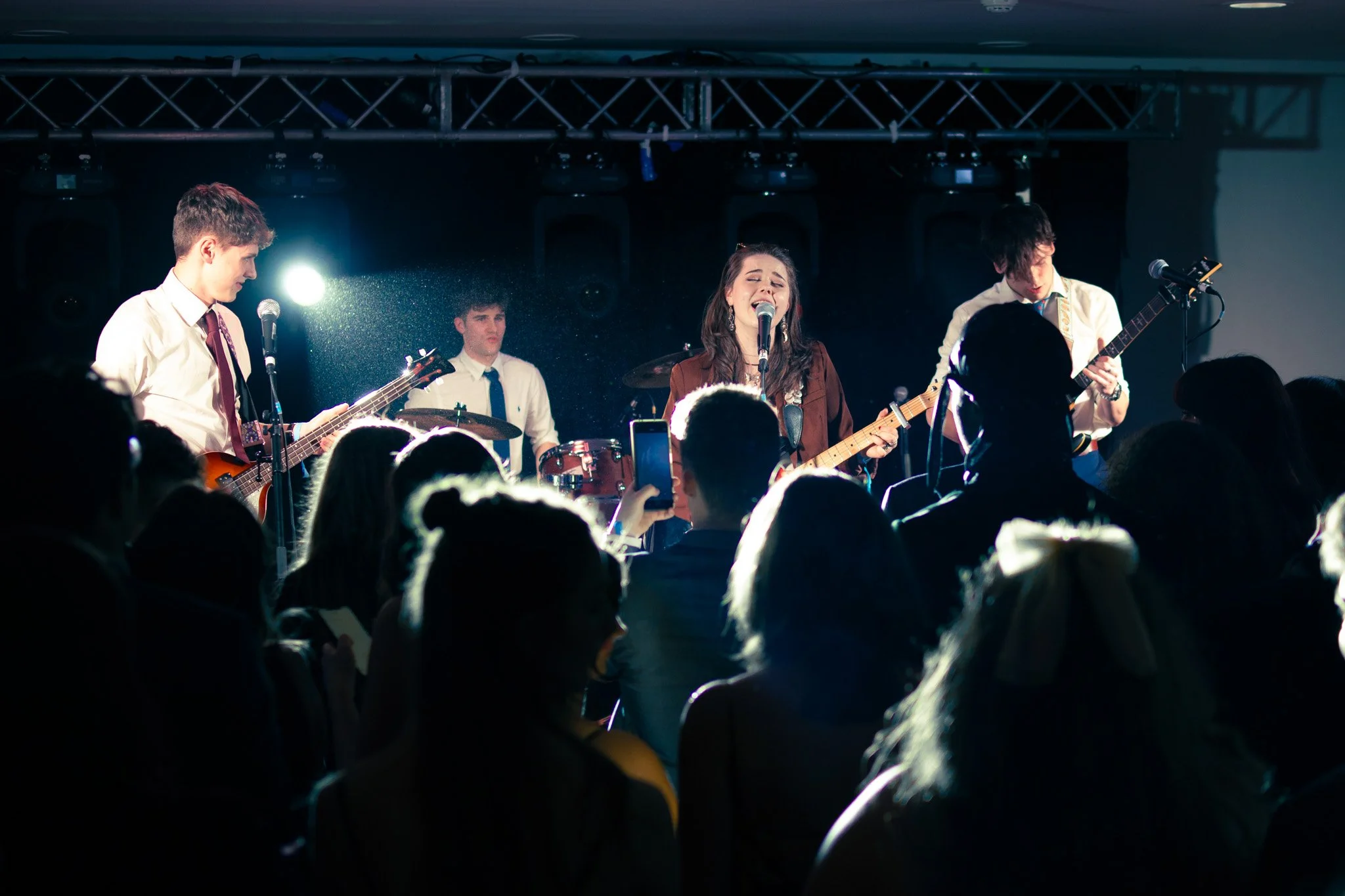 A band performing on stage with a female singer playing guitar, a male guitarist, a bassist, and a drummer, with an audience watching and some taking pictures.