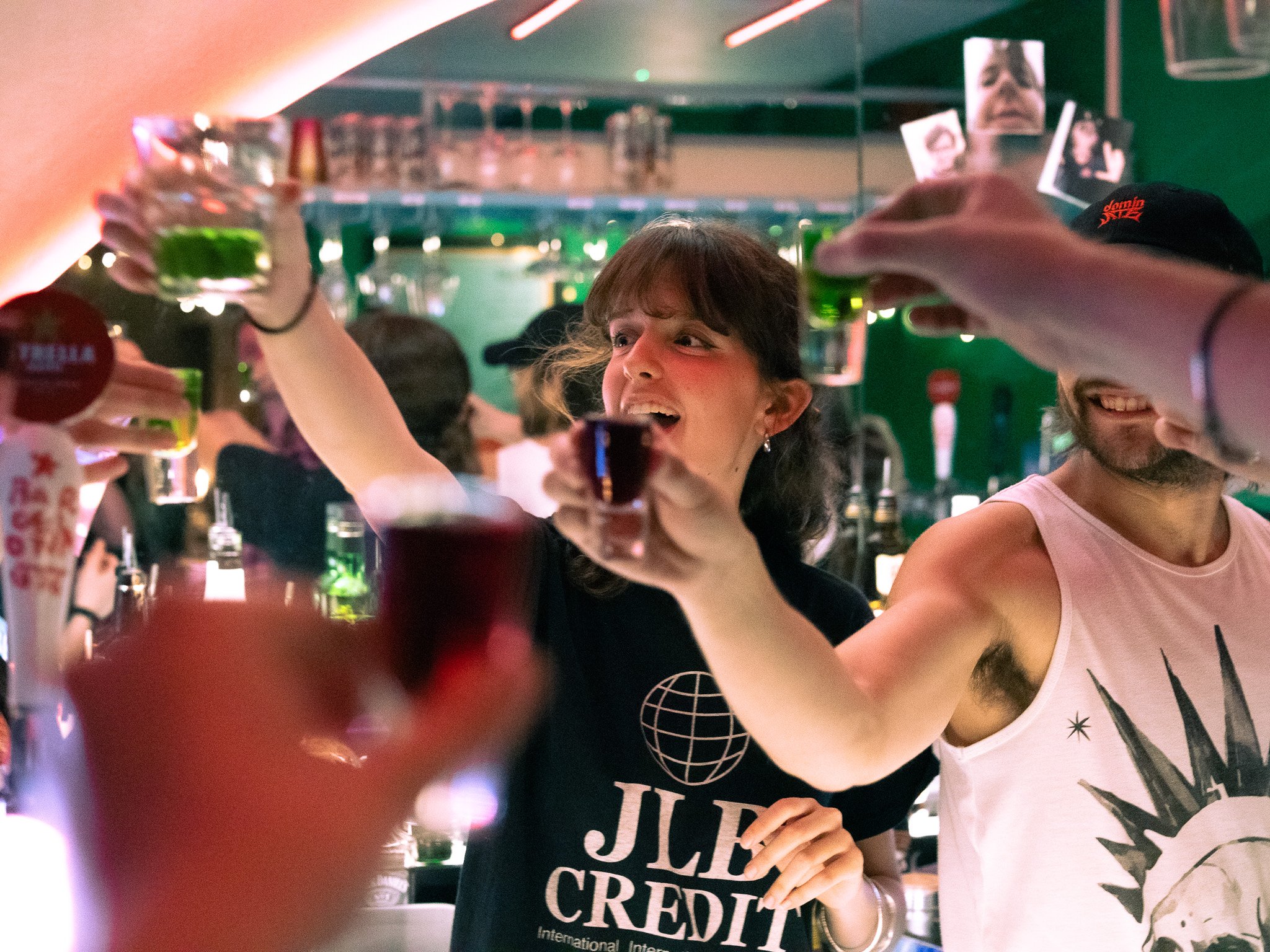 People in a bar raising shot glasses in a toast, enjoying drinks together. A woman with brown hair wearing a black shirt is at the center, smiling and reaching out with her glass. Next to her, a man with a beard and sleeveless shirt is also raising a