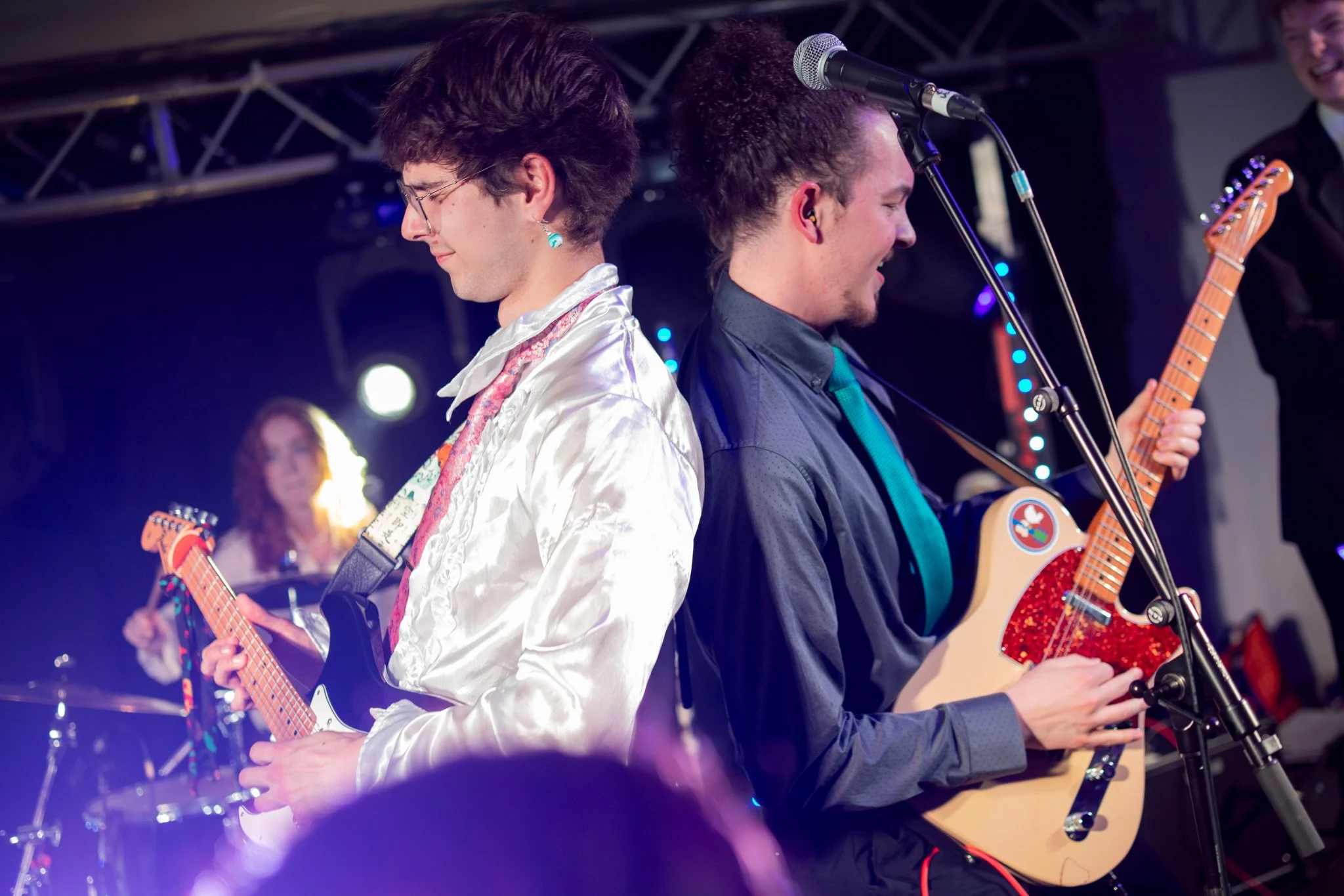 Two young men playing guitars back to back on stage during a live performance, with a woman in the background playing drums, all under stage lighting.