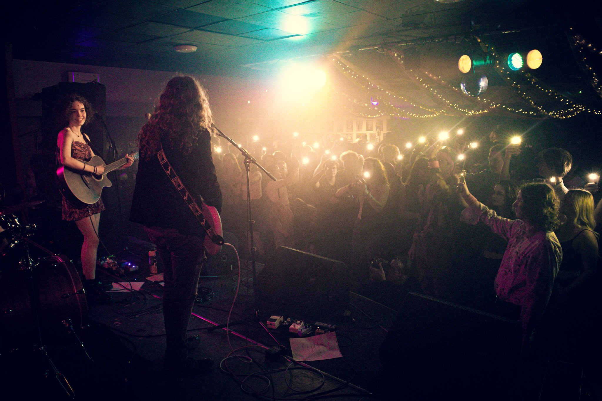 Two female musicians perform on stage with guitars in front of an audience in a dimly lit venue decorated with string lights. The crowd is enthusiastic, with some people capturing the moment with their phones.