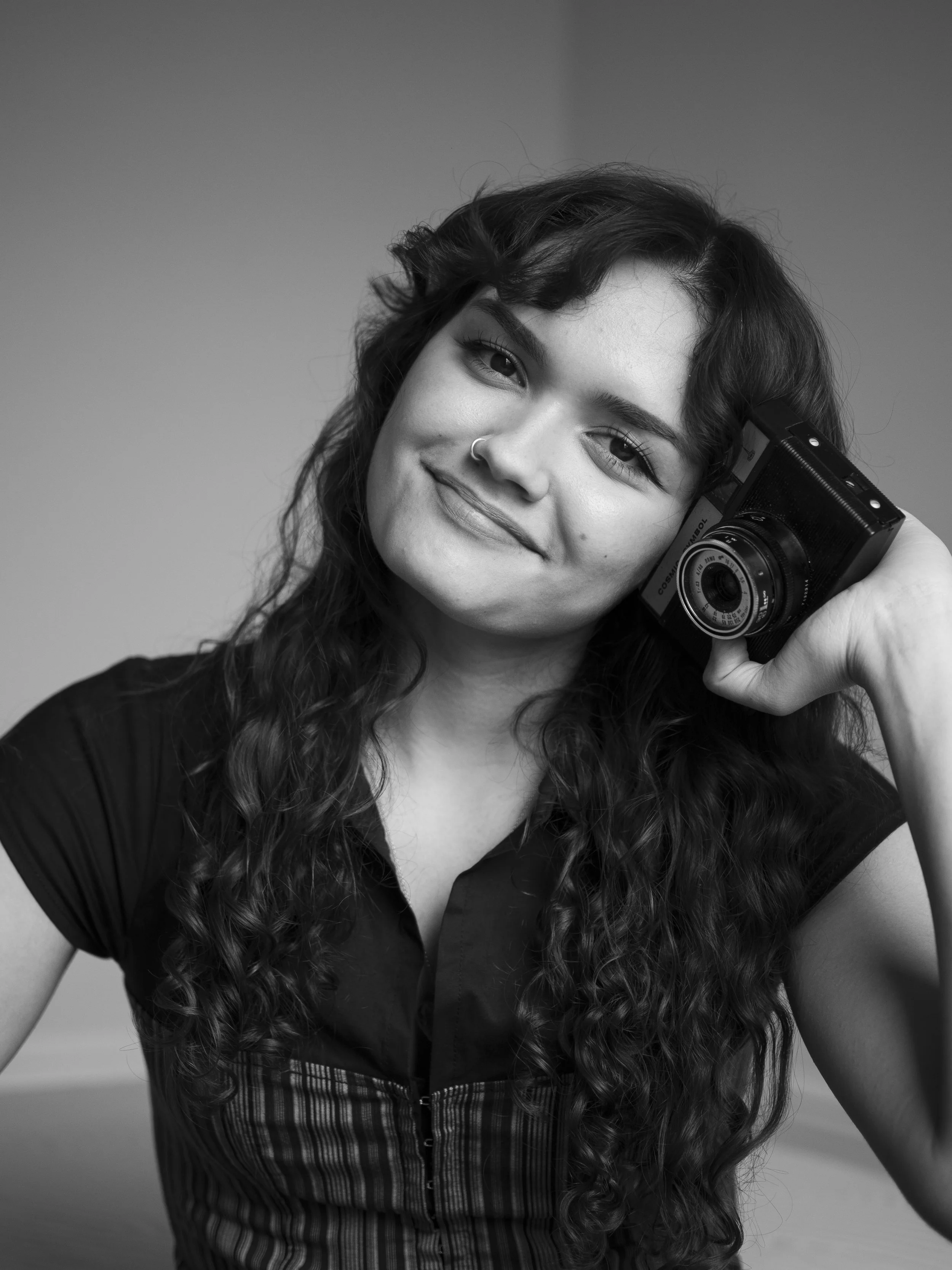 A young woman with curly hair and a nose ring smiling while holding a vintage camera to her ear.
