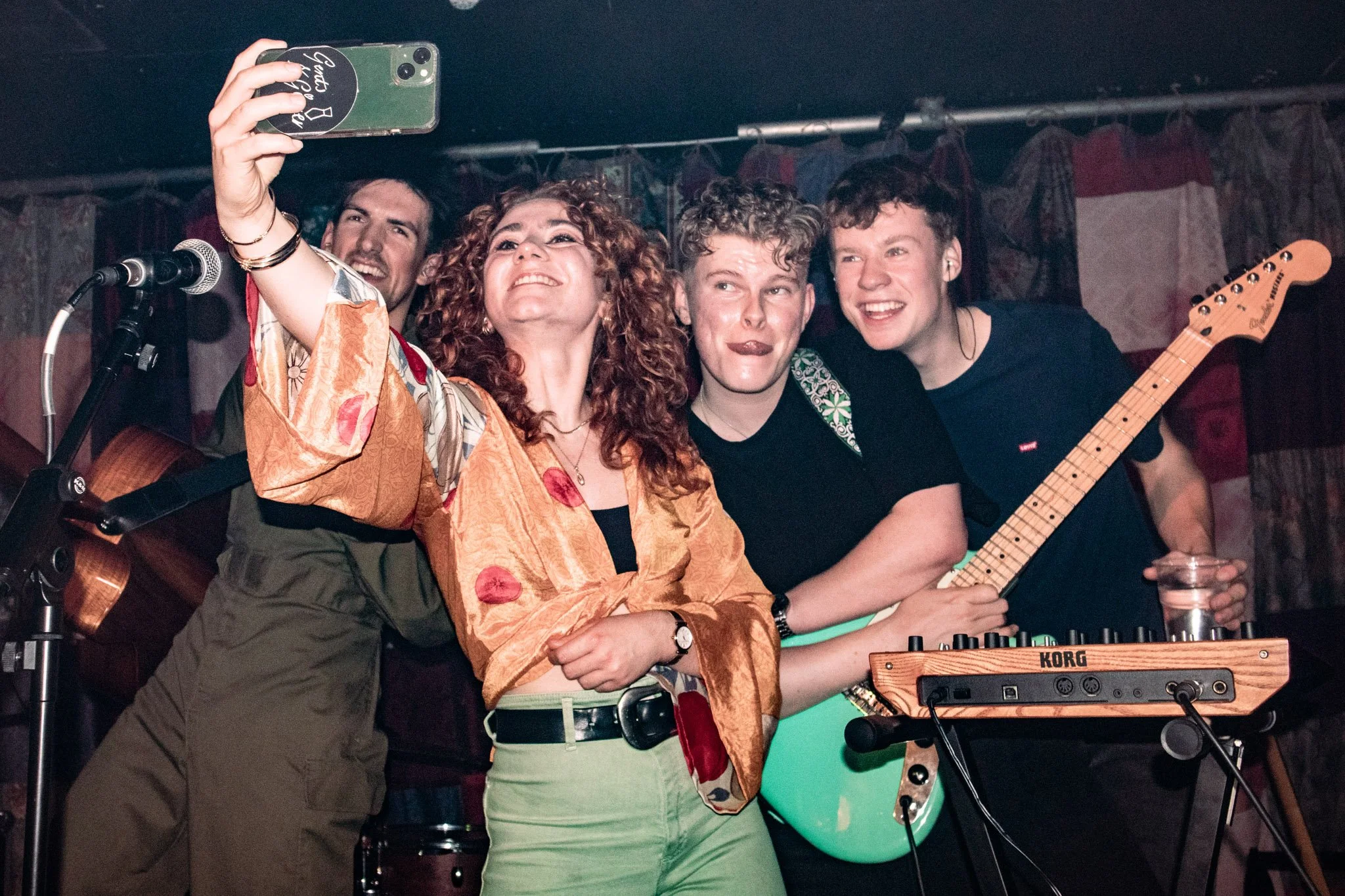 Four people taking a selfie on stage with musical instruments and microphone in a dimly lit venue.