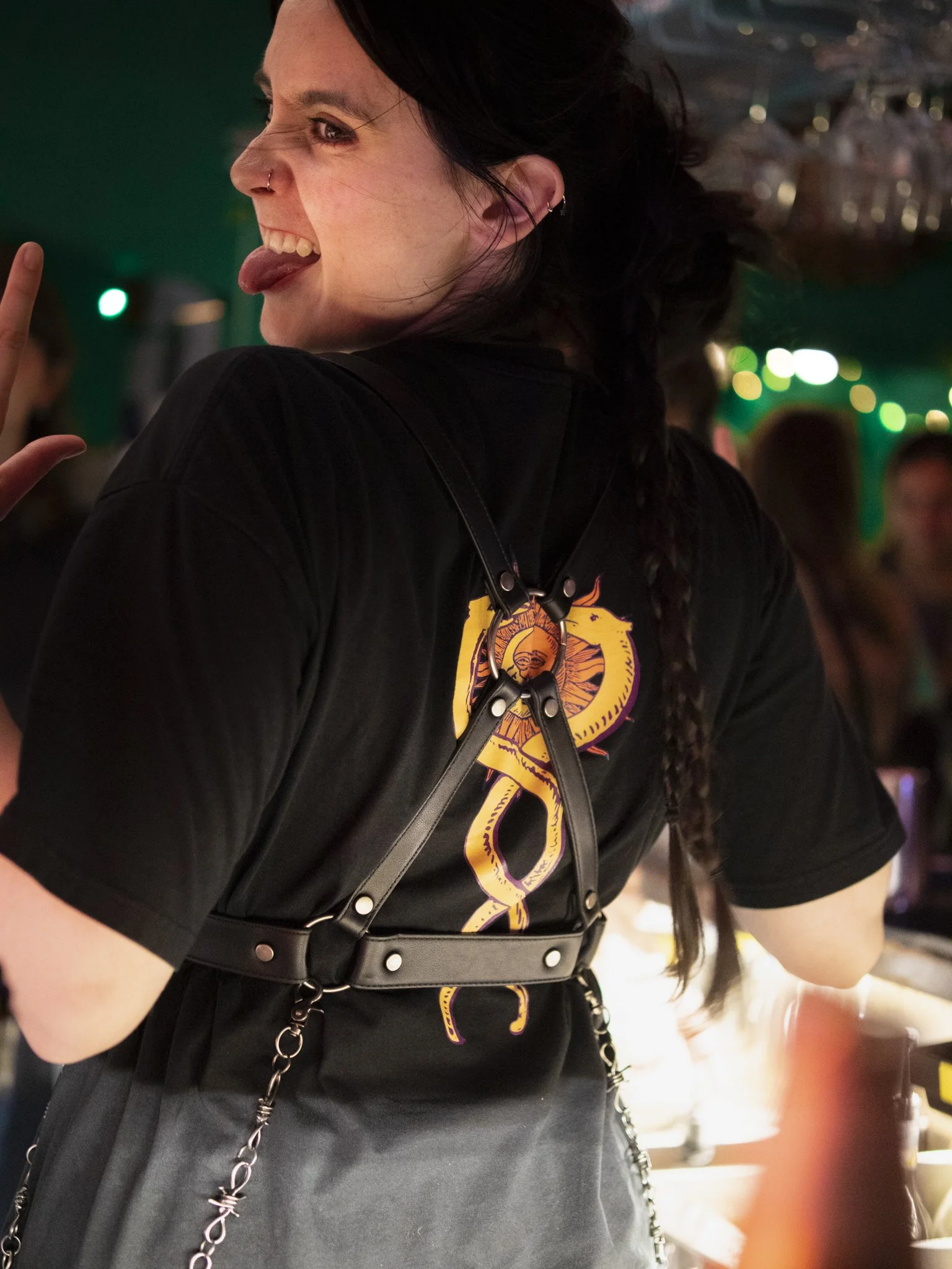A woman with dark hair in braids, wearing a black t-shirt with a yellow and purple design, making a playful face with her tongue out, at a lively indoor event with colorful lights and other people in the background.