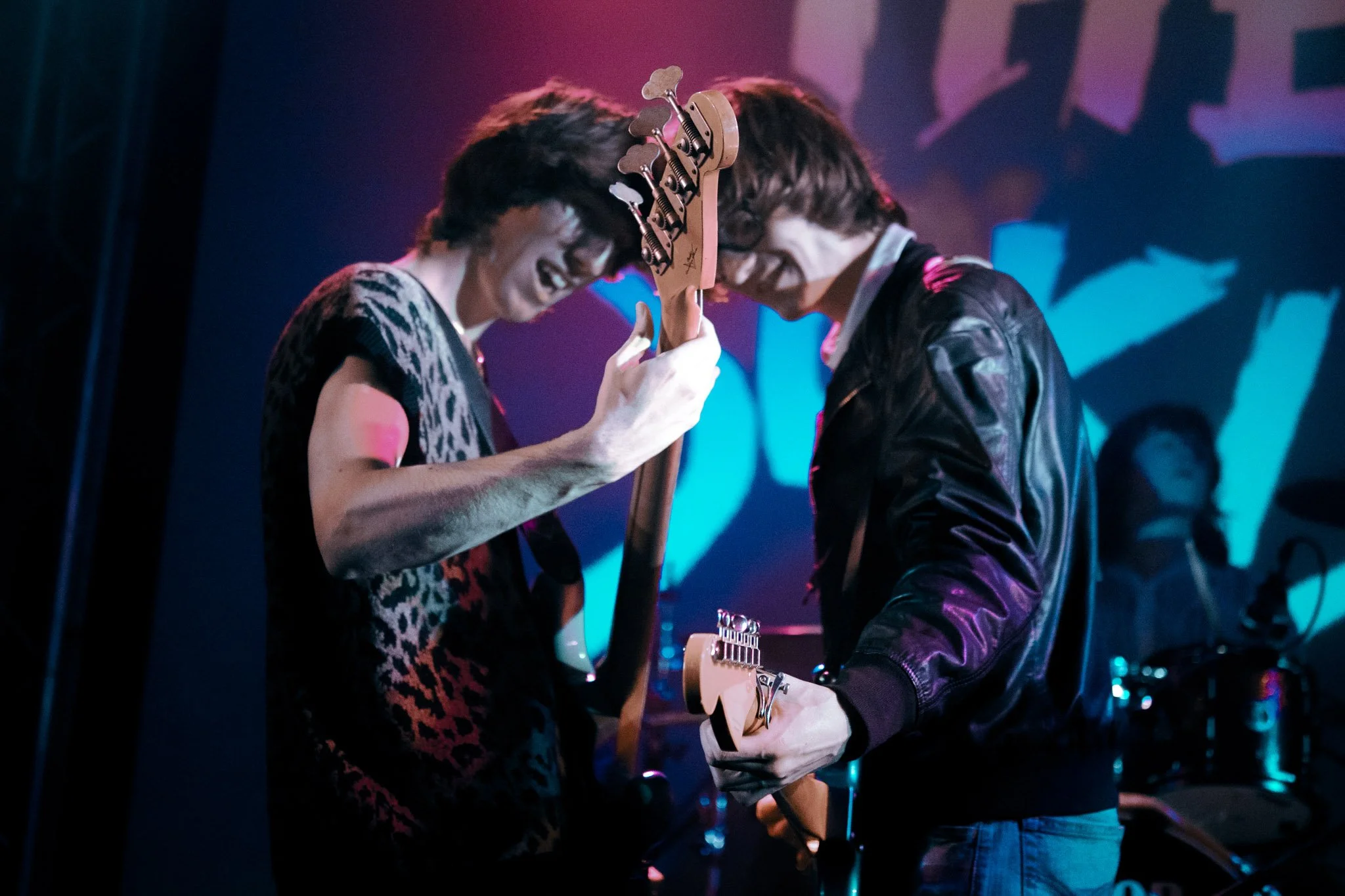 Two musicians with long hair, wearing dark clothing, are playing electric guitars face-to-face on stage with a drummer visible in the background. The stage is lit with colorful lights.