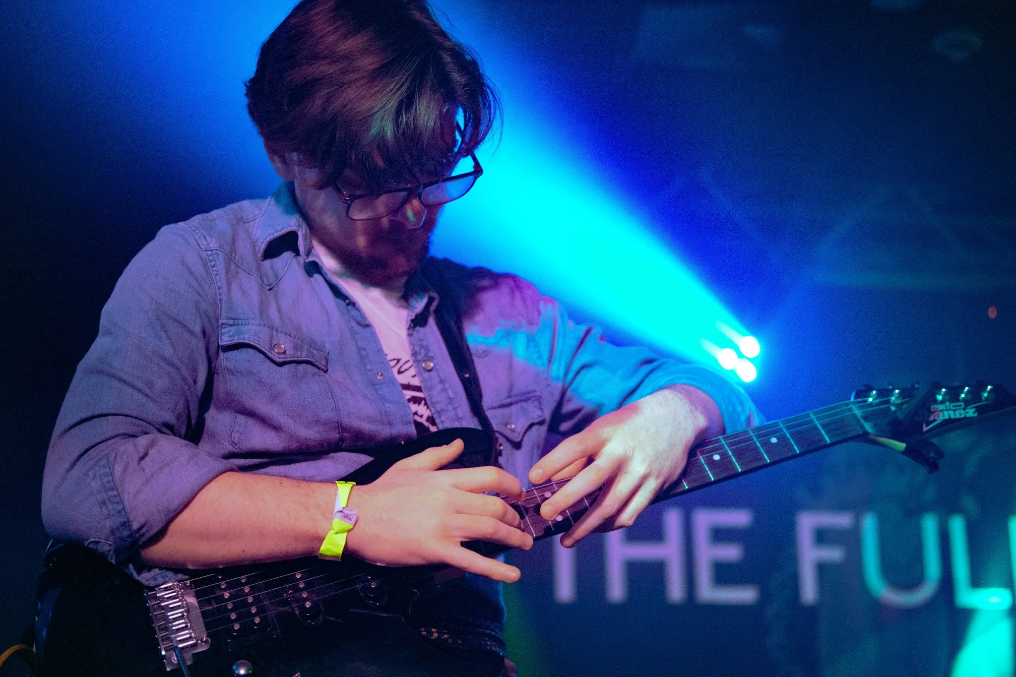 A man with curly hair and glasses playing an electric guitar on stage, illuminated by blue stage lights.