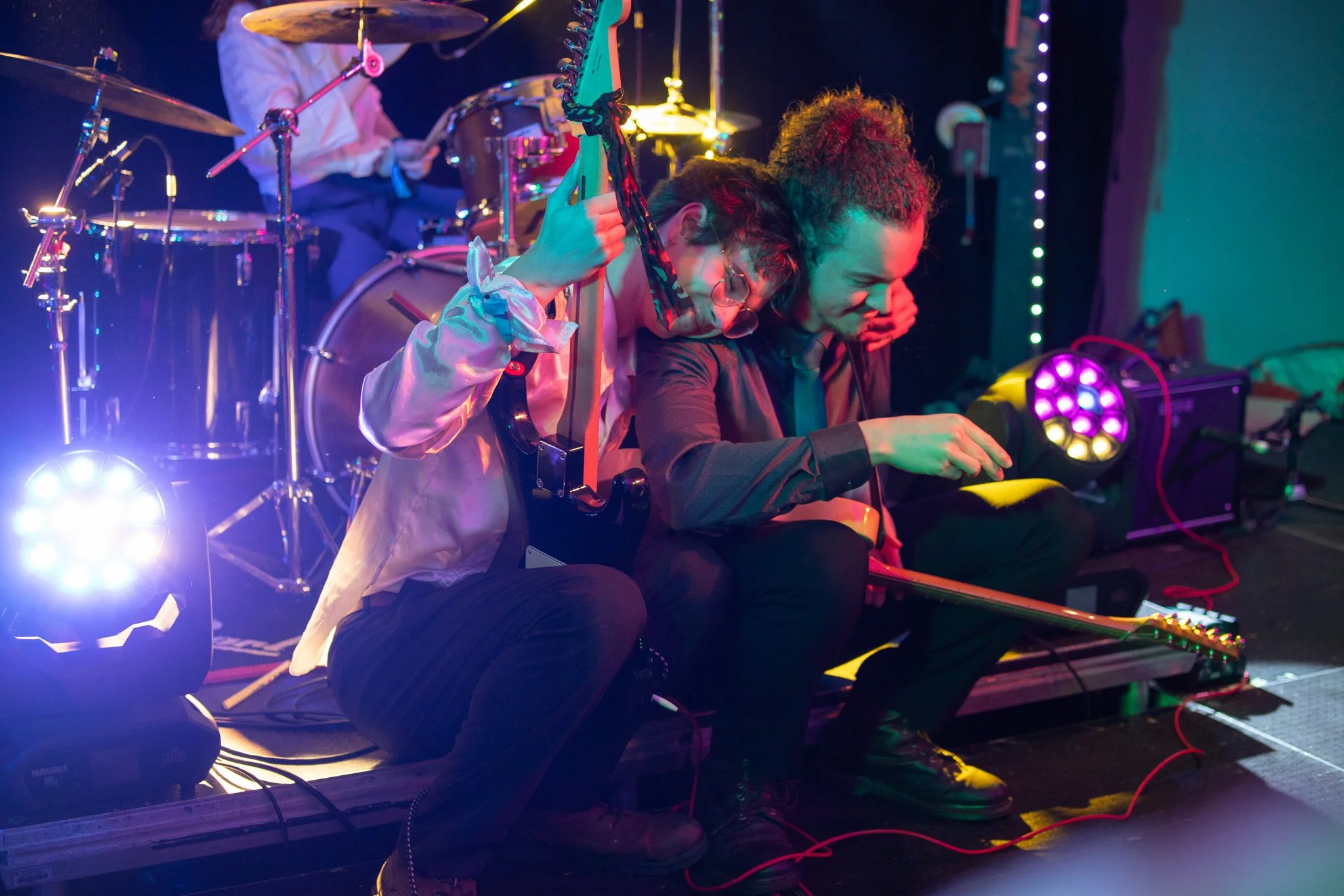 Two musicians seated on the stage, one adjusting an electric guitar and the other playing an electric guitar, with a drum set and stage lights in the background.