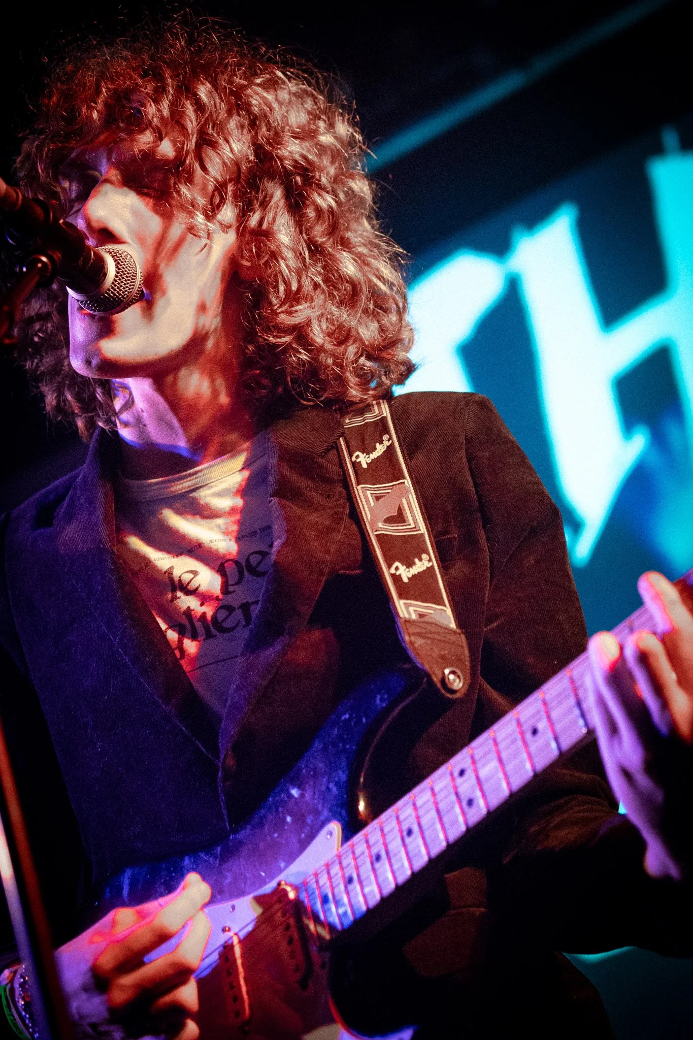 A musician with curly hair singing into a microphone and playing an electric guitar during a live performance, with stage lighting and a screen in the background.