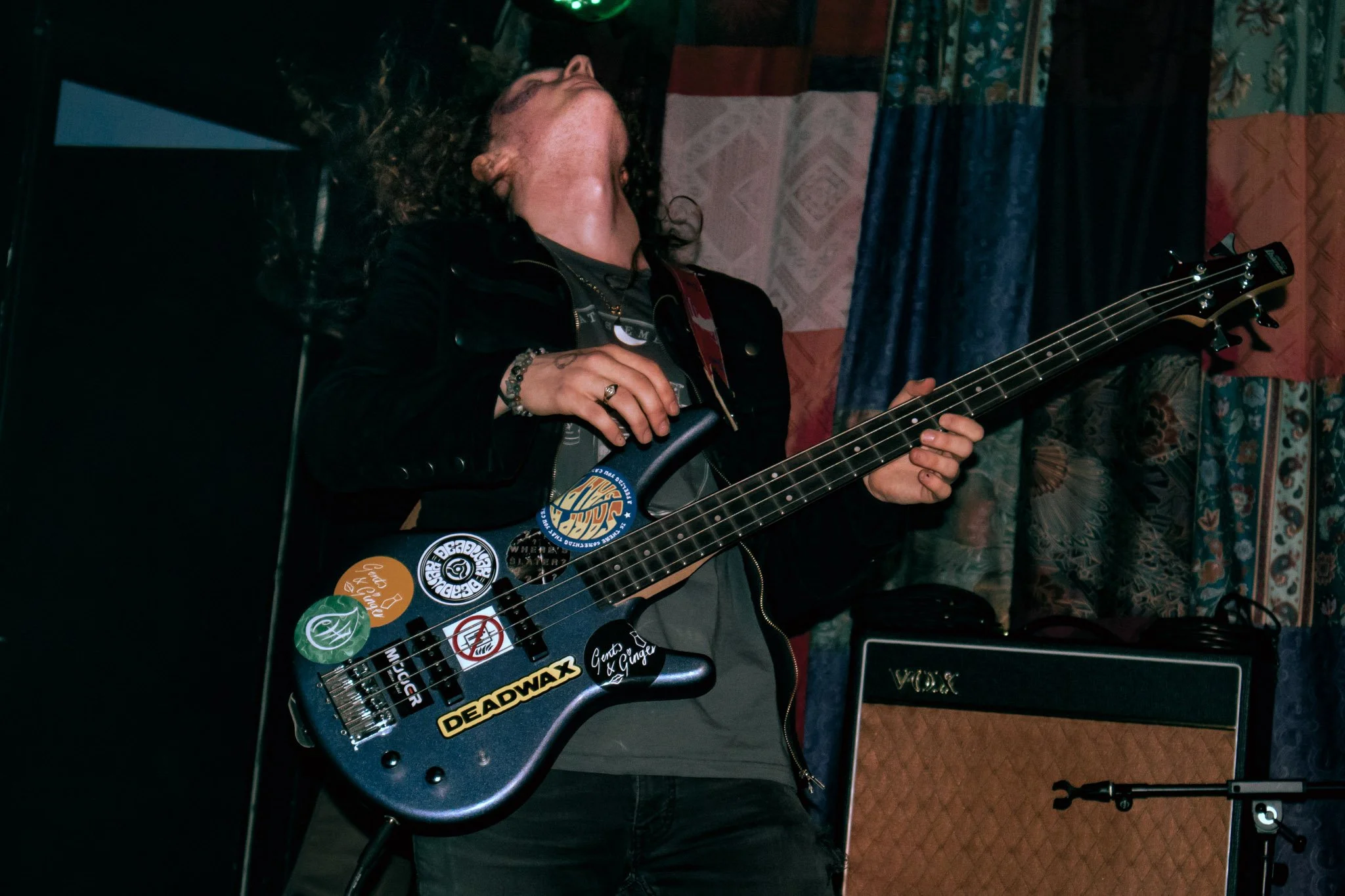 A person with long curly hair playing an electric guitar covered with stickers, with a Vox amplifier in the background, indoors with patterned curtains.