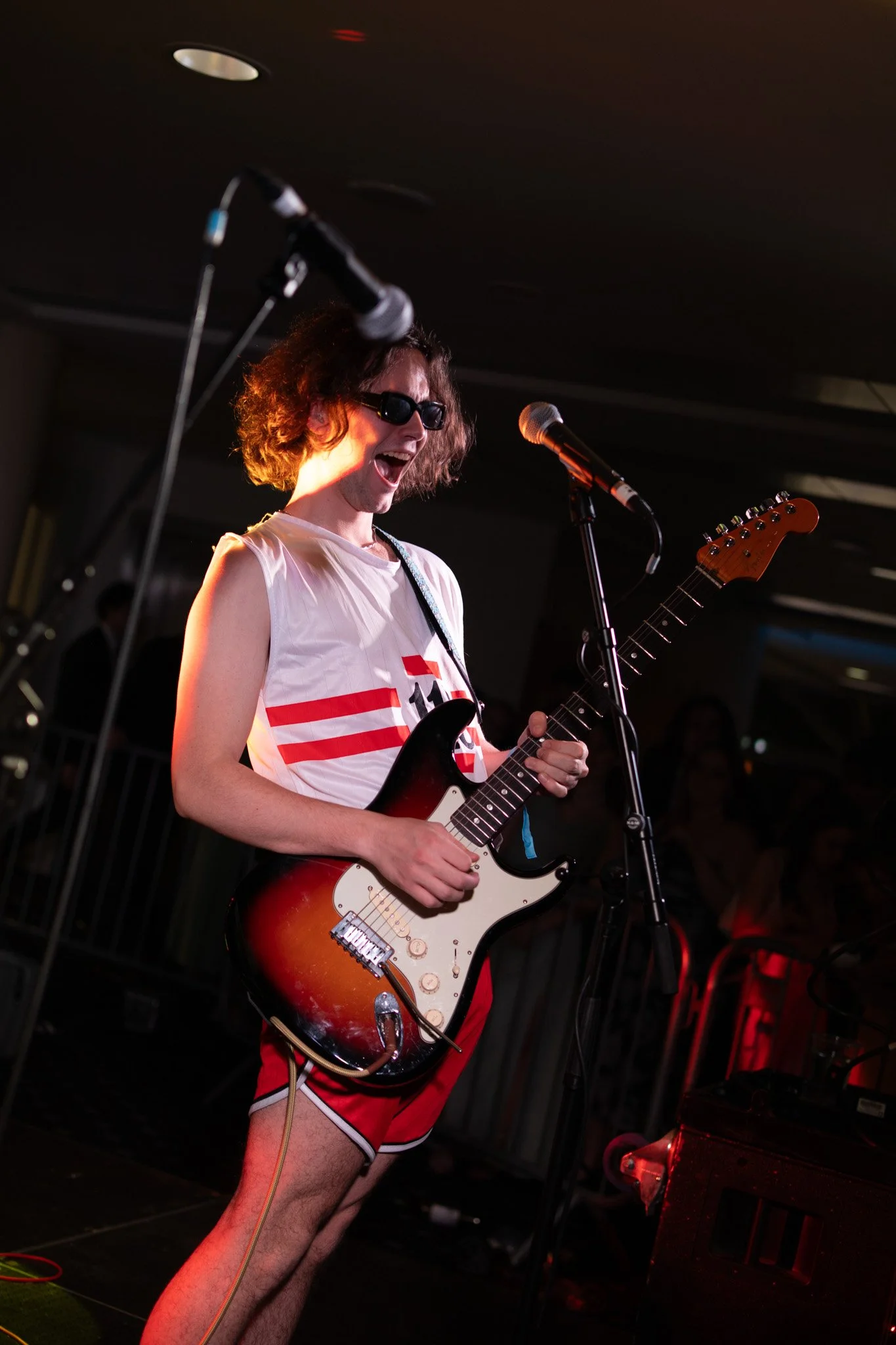 Person playing electric guitar on stage, wearing sunglasses, sleeveless shirt, and red shorts, live music performance.