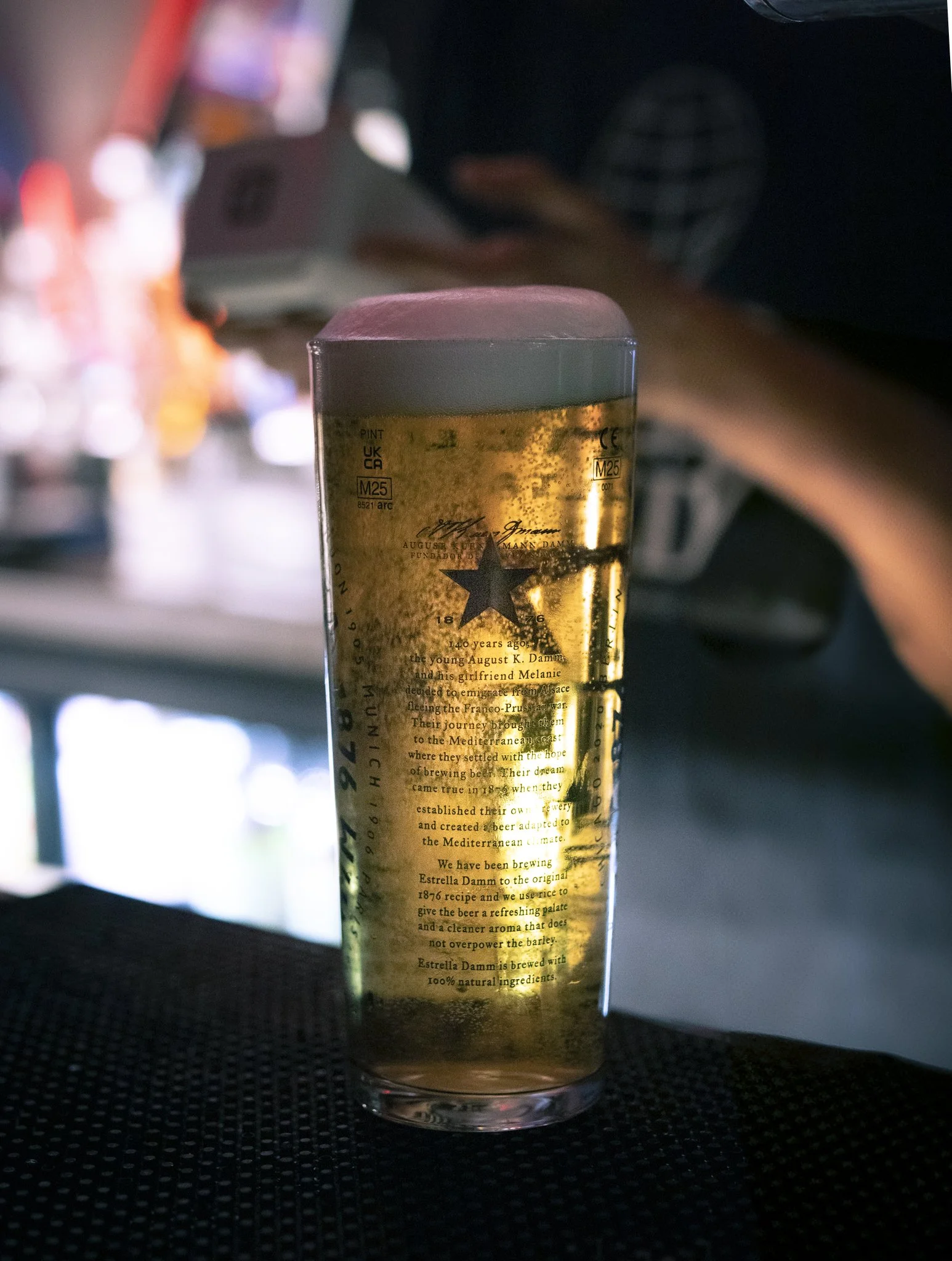 A tall glass of beer with a frothy head, labeled with a star logo and text, filled with golden beer, placed on a dark surface. Background shows a person using a smartphone with a blurred indoor setting.
