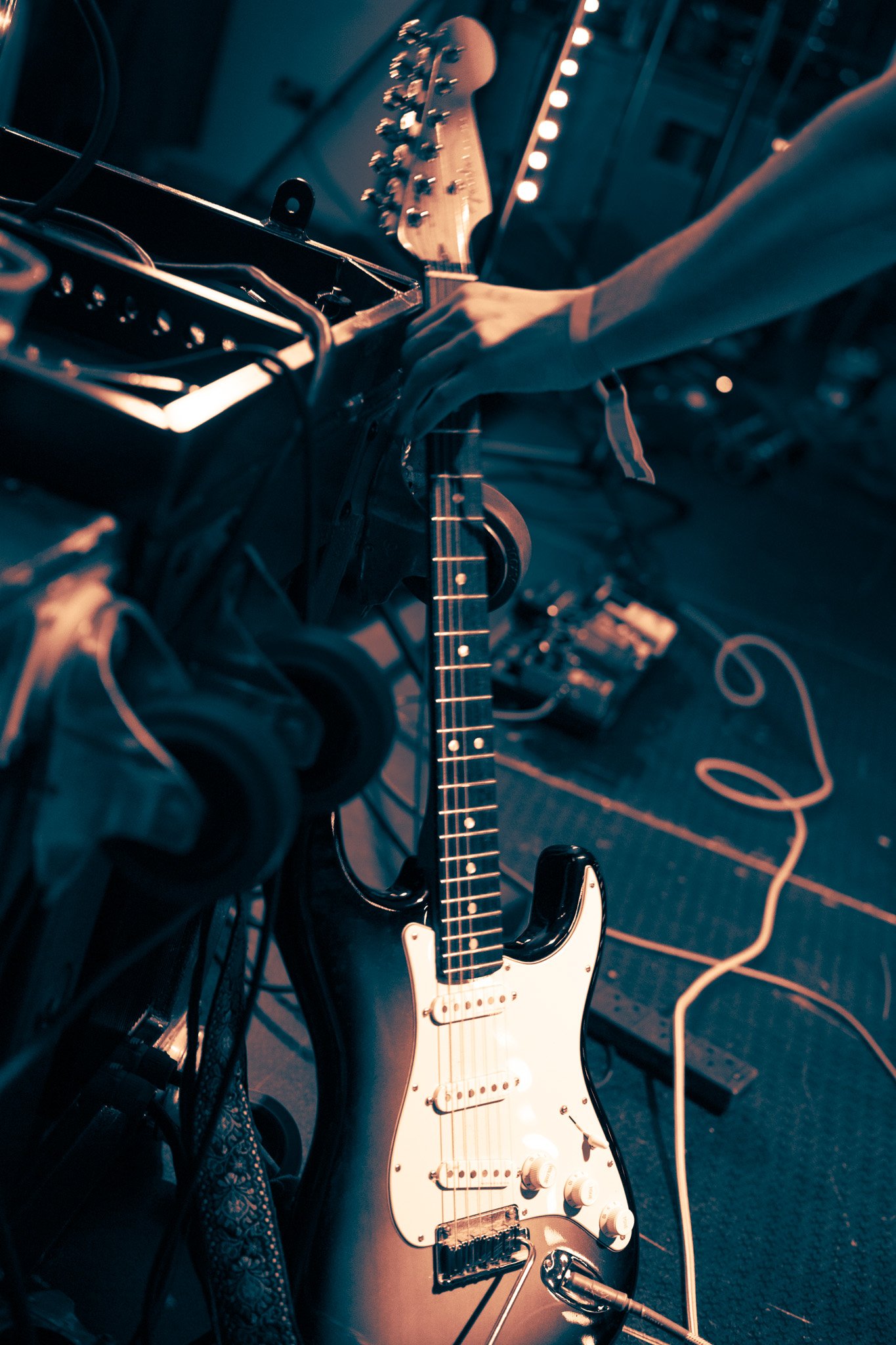 Electric guitar leaning against equipment on stage with a person's arm reaching for it, cord on the floor, and strings of lights in the background.