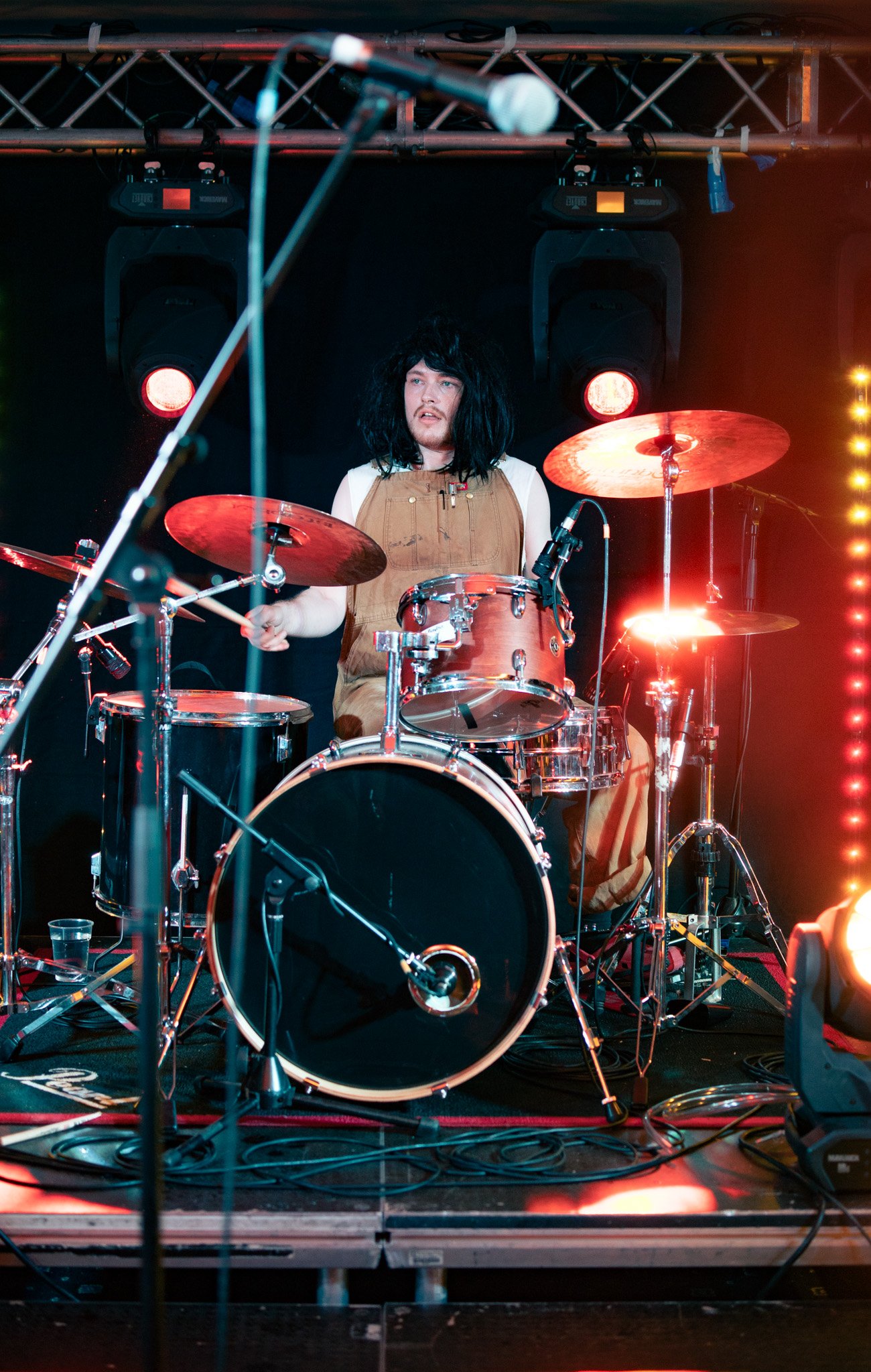 A person with black, curly hair wearing a tan jumpsuit is playing a drum set on a stage with red and yellow lights.