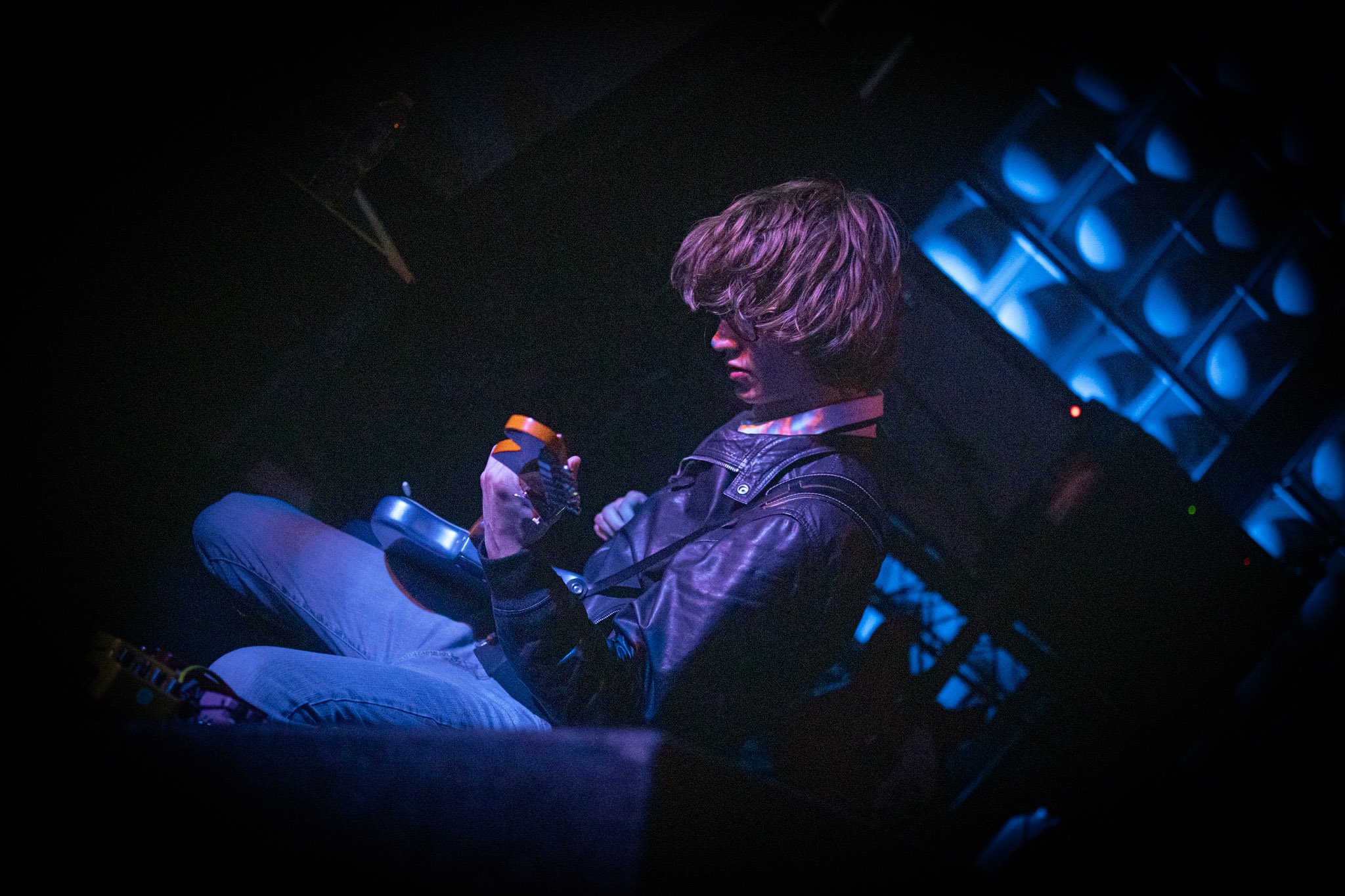 Person with wavy hair sitting in a dark room, holding a game controller and looking down, illuminated by blue lighting.