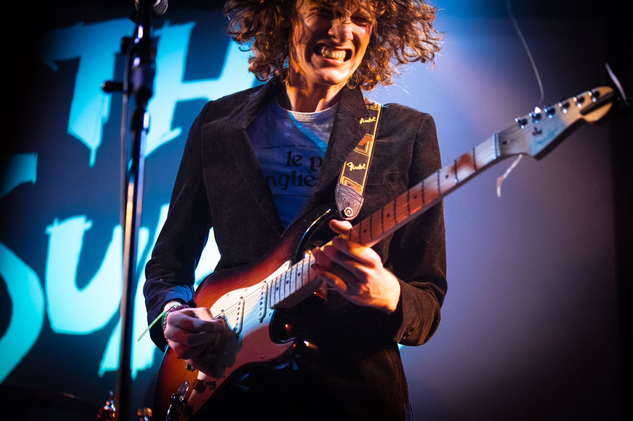 A person with curly hair playing an electric guitar on stage, wearing a black jacket and a t-shirt, with a Fender guitar strap, in front of a backdrop with blue lighting.