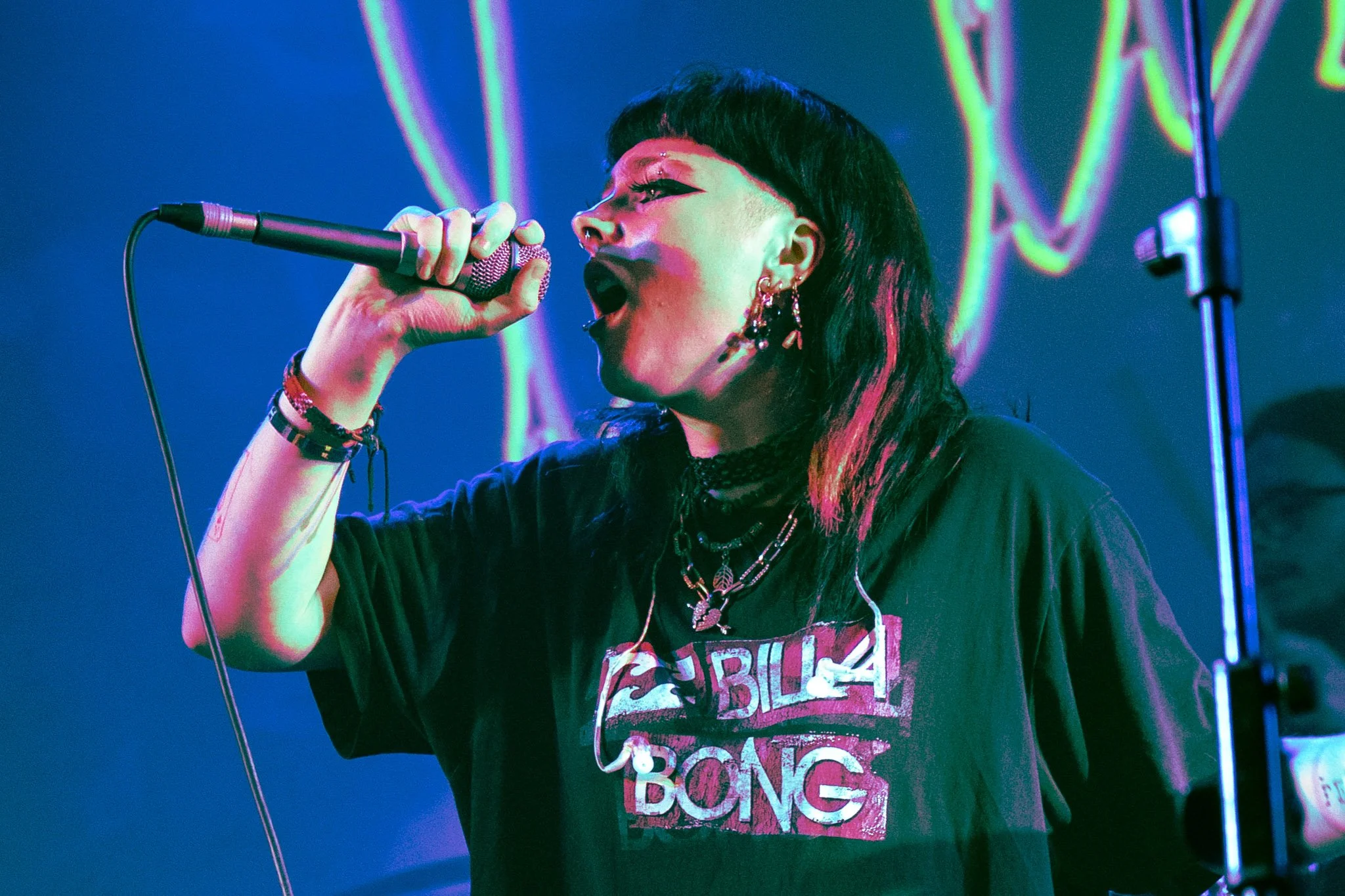 A woman with dark hair singing into a microphone on stage with colorful neon lights in the background.