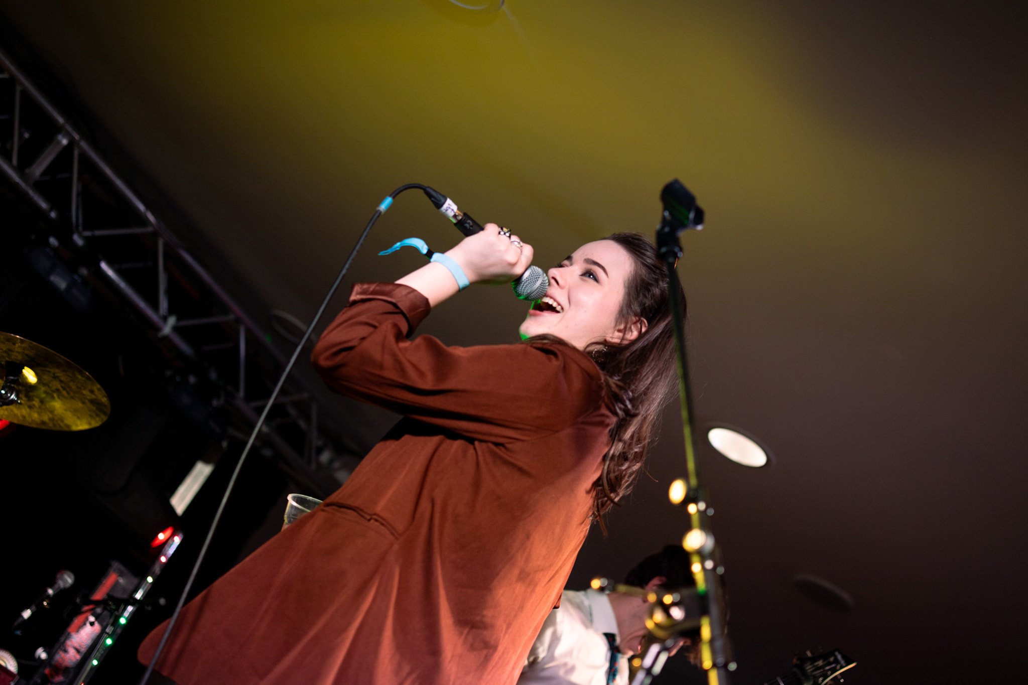A woman singing into a microphone on stage, wearing a brown jacket, with stage lighting and equipment visible in the background.