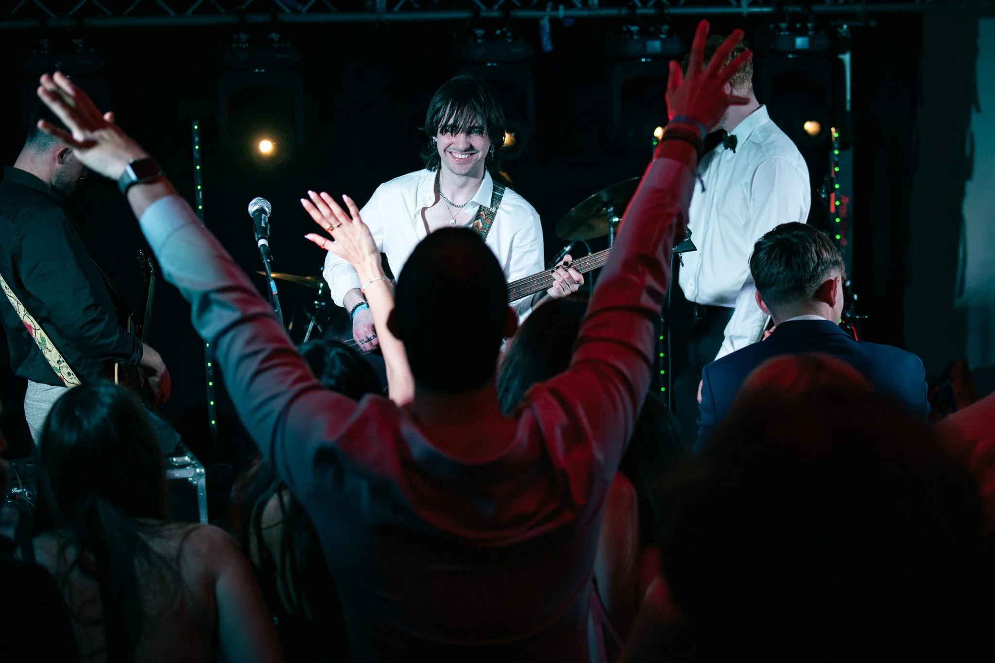 A lively crowd at a concert with a band performing on stage; one musician with long dark hair, smiling and playing an electric guitar, wearing a white shirt. Several audience members have their hands raised in the air.