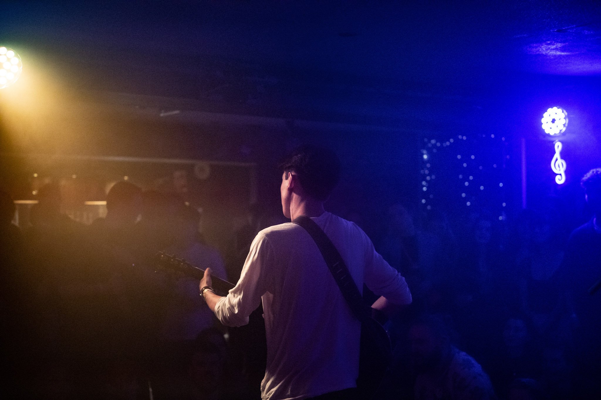 Person playing guitar on stage with blue and yellow stage lights, audience in background, decorative lighting and music note sign on wall.