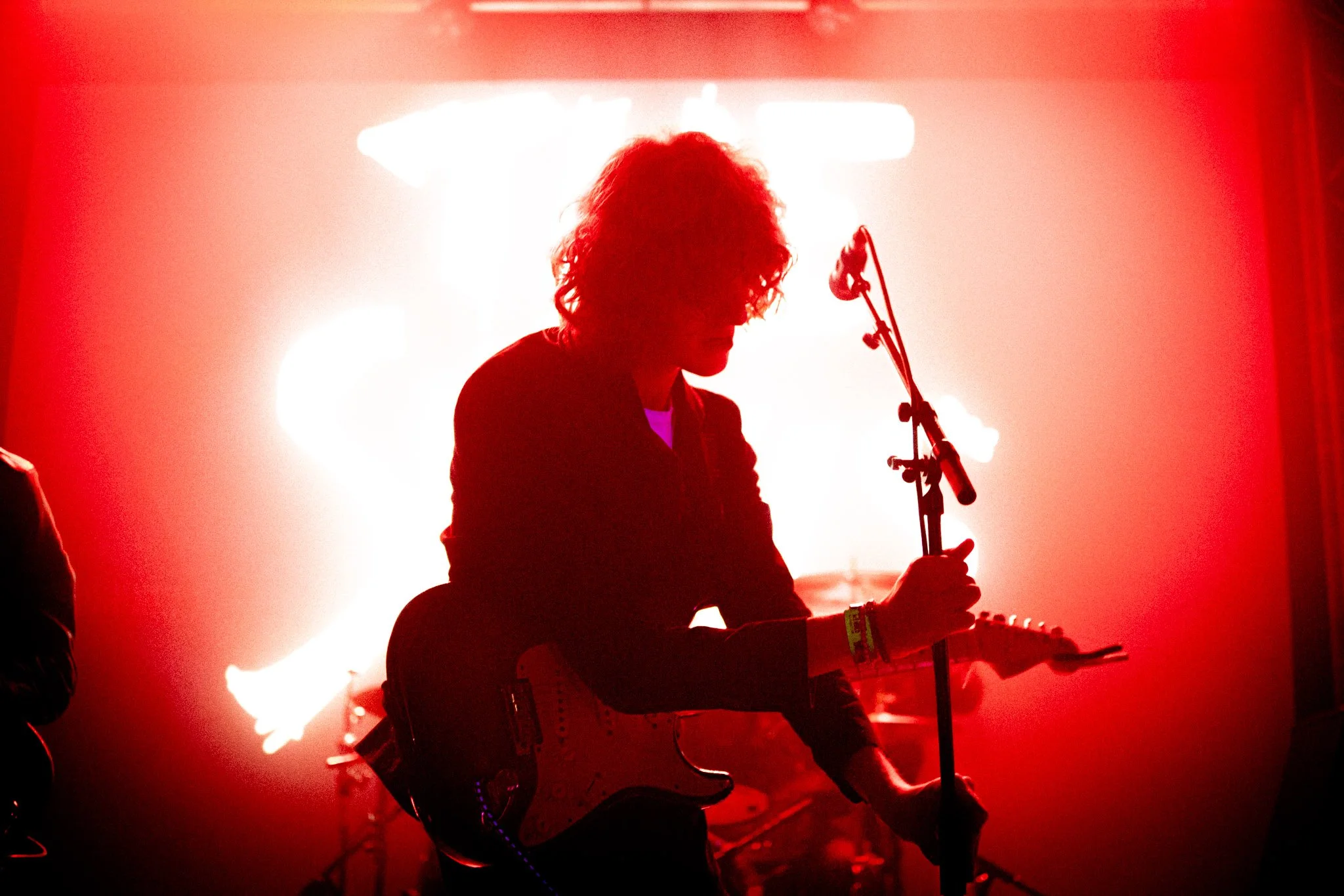 A silhouette of a person with curly hair playing an electric guitar on stage with bright backlighting and red hues.