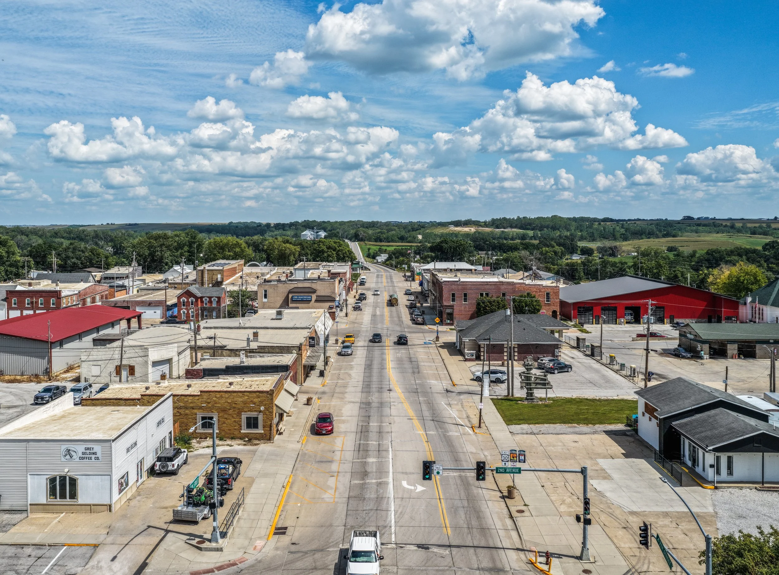 A small town street scene under a partly cloudy sky with green hills in the background. The street is lined with buildings, parked cars, and a traffic signal at the intersection, labeled as 2nd Avenue and East 30.