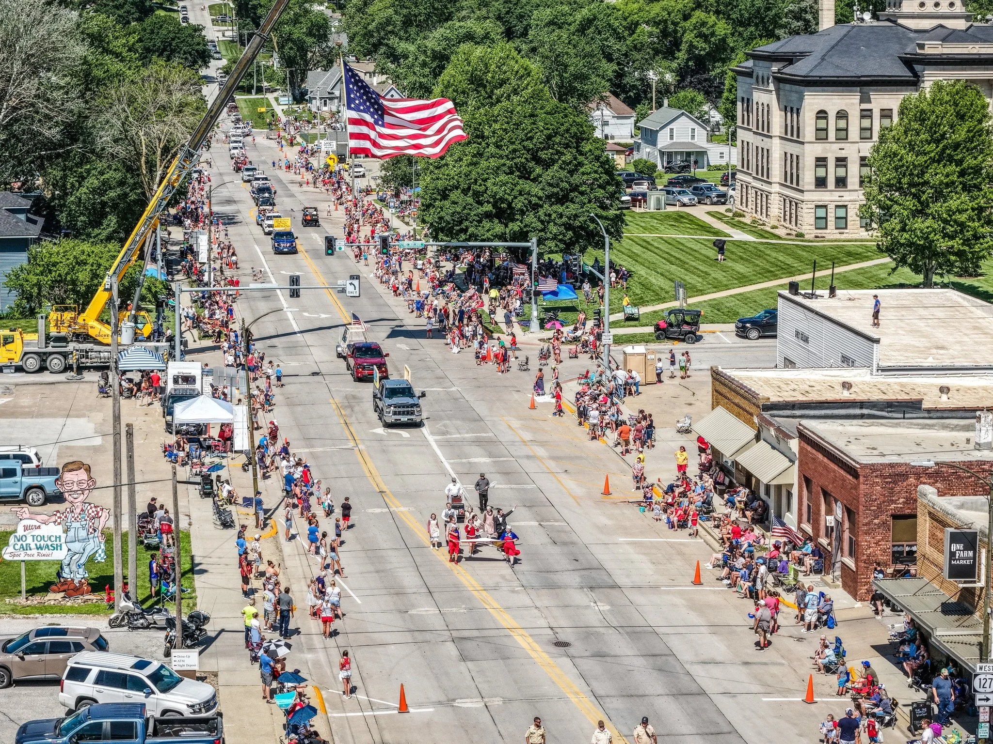 Logan, Iowa's annual 4th of July parade, with an American flag on a crane leading parade participants. Crowds of people line the streets and there are cars parked along the street and tents set up on the sidewalks.