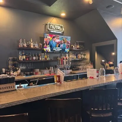 Taproom 1890's bar with liquor bottles, a TV showing a talk show and a neon sign reading 'NUTRL'.