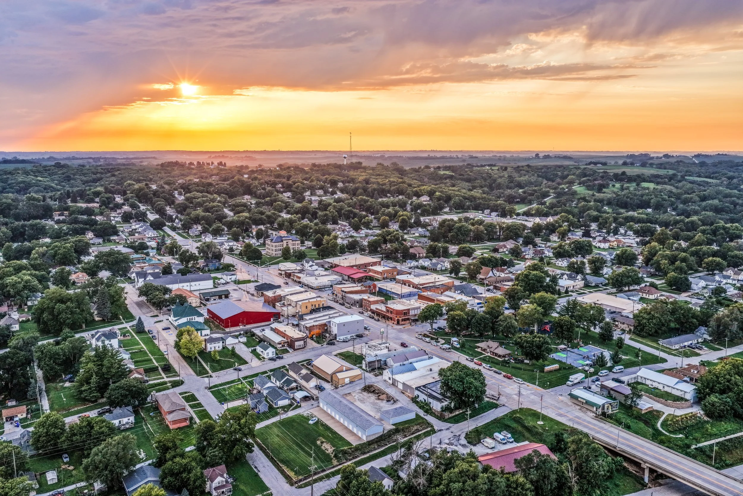 Aerial view of small-town Logan during sunset with trees, residential houses, commercial buildings and a few sports courts visible.