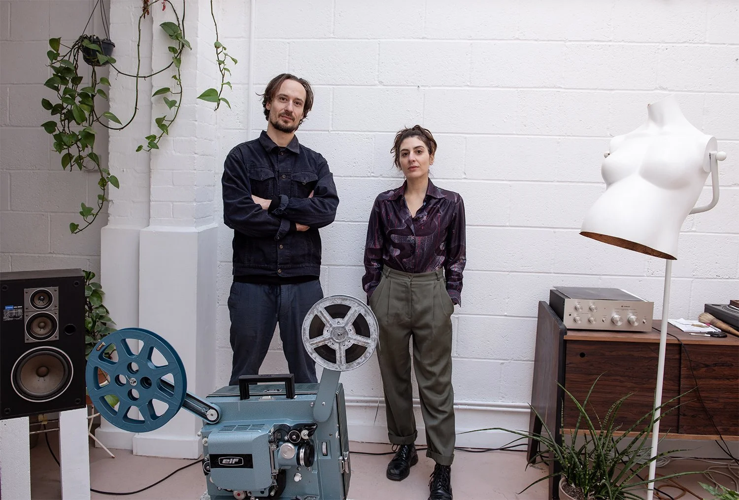 A man and a woman in a converted warehouse studio in London, looking at the camera amongst objects they have made and objects that they use to work with in their creative studio. There is a hi-fi set up, a 16mm projector, and handmade furniture