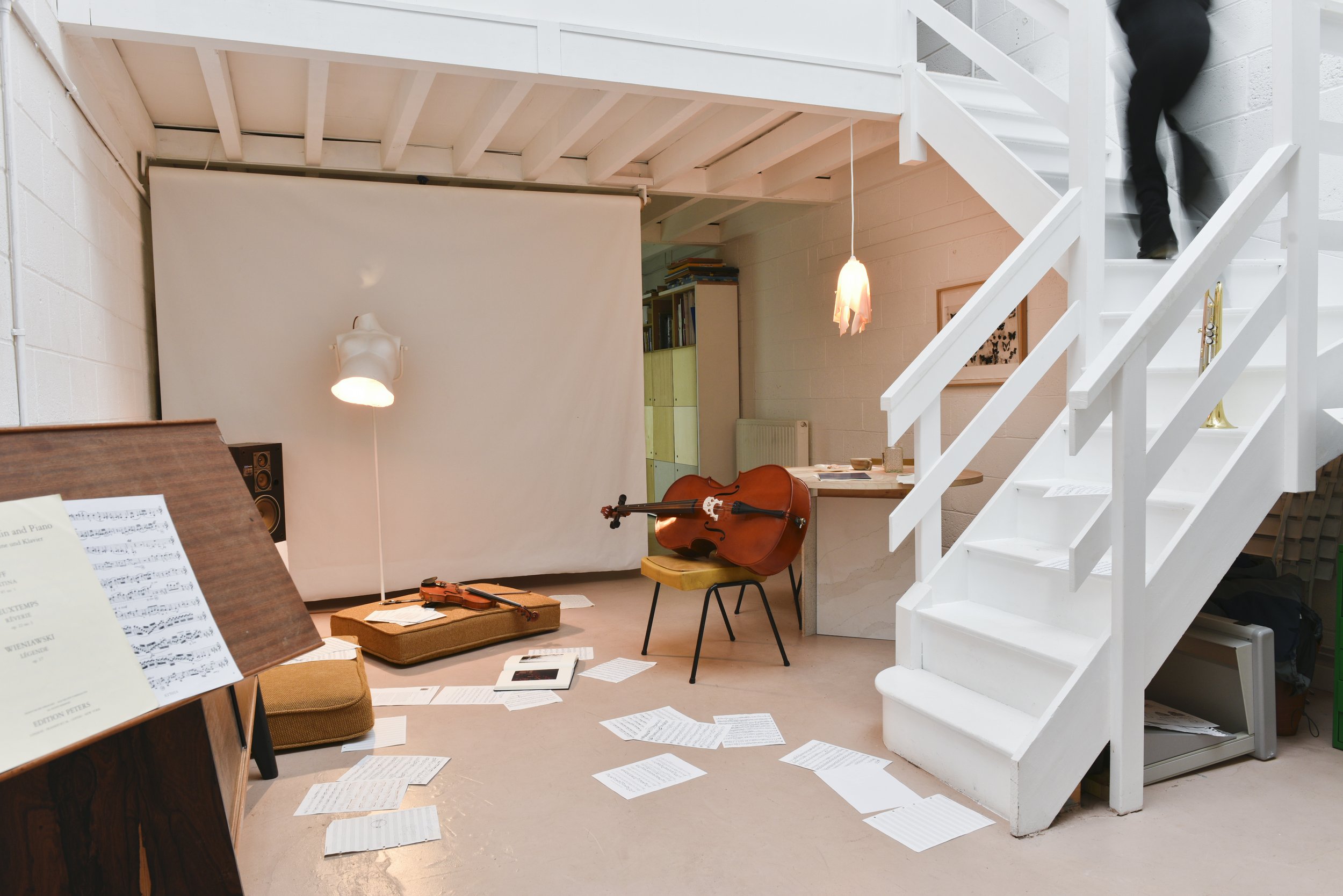 A space with music dispersed on the floor and a light on with someone who must be in the middle of working on a music score. There is a cello resting on a seat in the middle of the space and someone is walking up the stairs