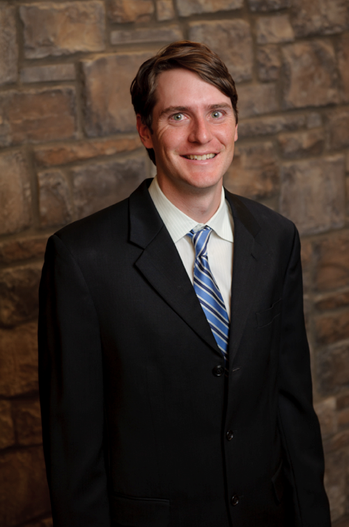Young man in a black suit with a striped blue tie, smiling, standing in front of a brick wall.