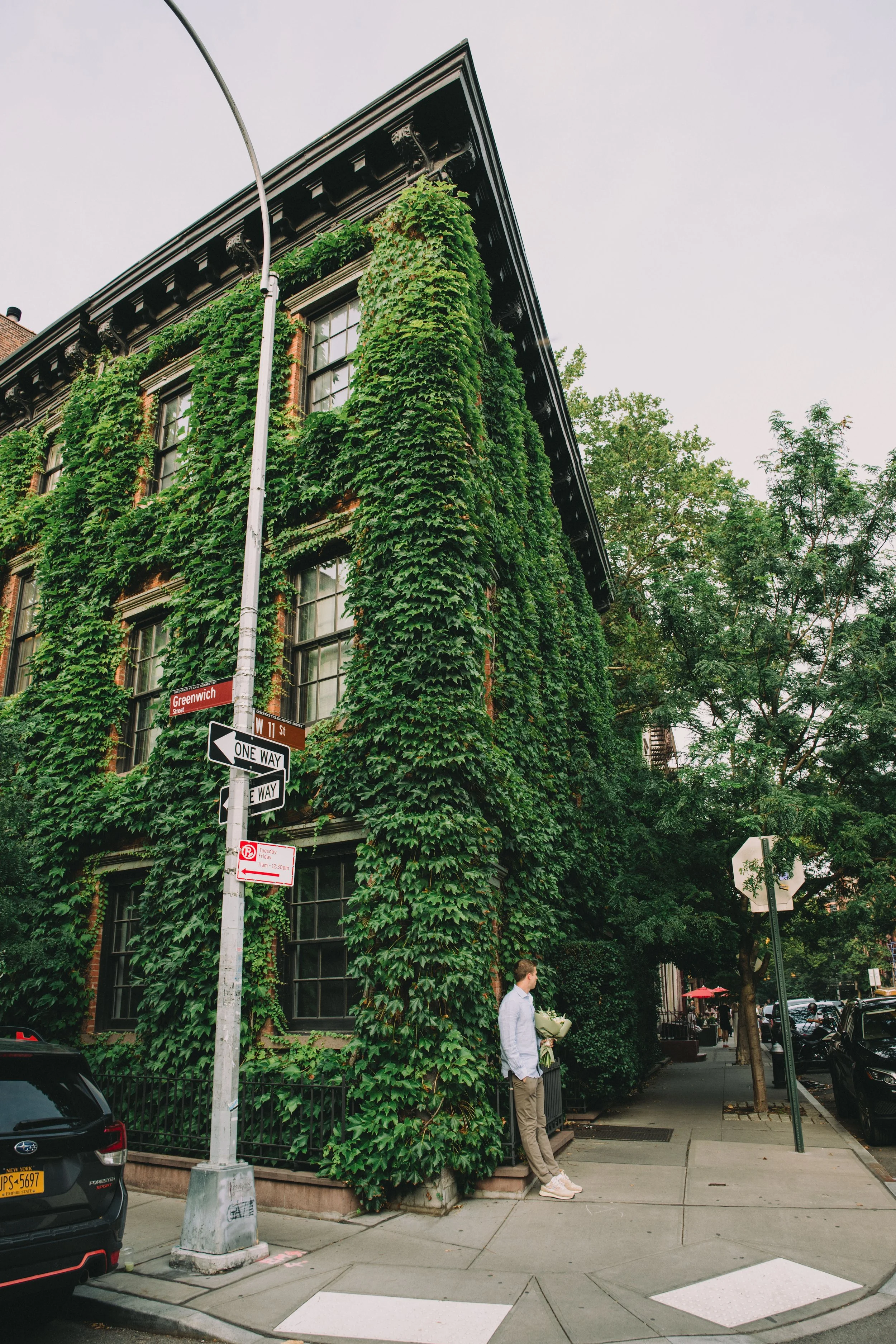 A man standing on a city sidewalk next to a building covered in green ivy, with street signs indicating Greenwich Street and West 11th Street, and a streetlight in the foreground.