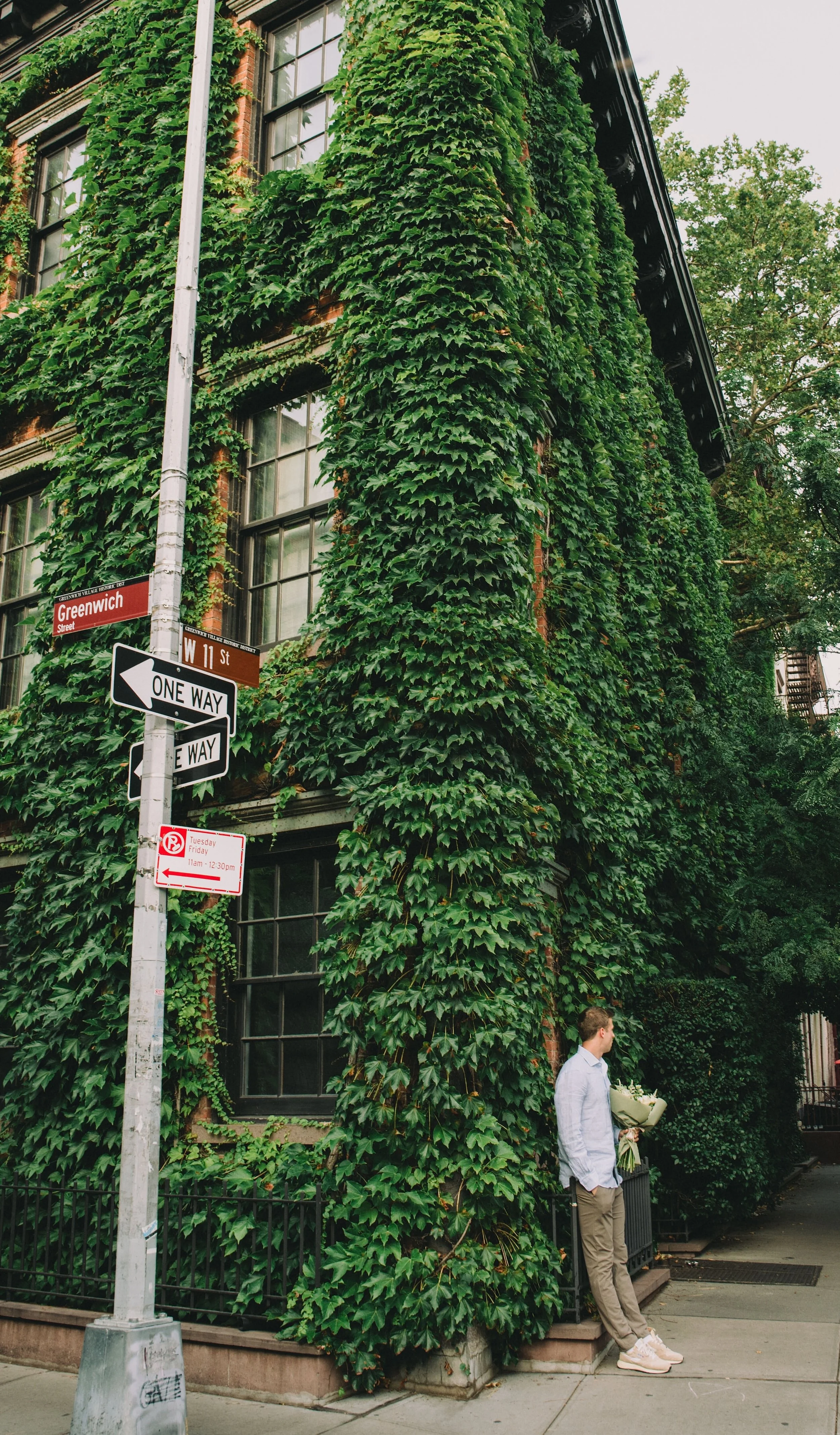 A man standing on a sidewalk next to a building covered in green ivy, holding a bouquet of flowers, with street signs indicating Greenwich Street and W 11th Street, and various traffic and parking signs, during the daytime.