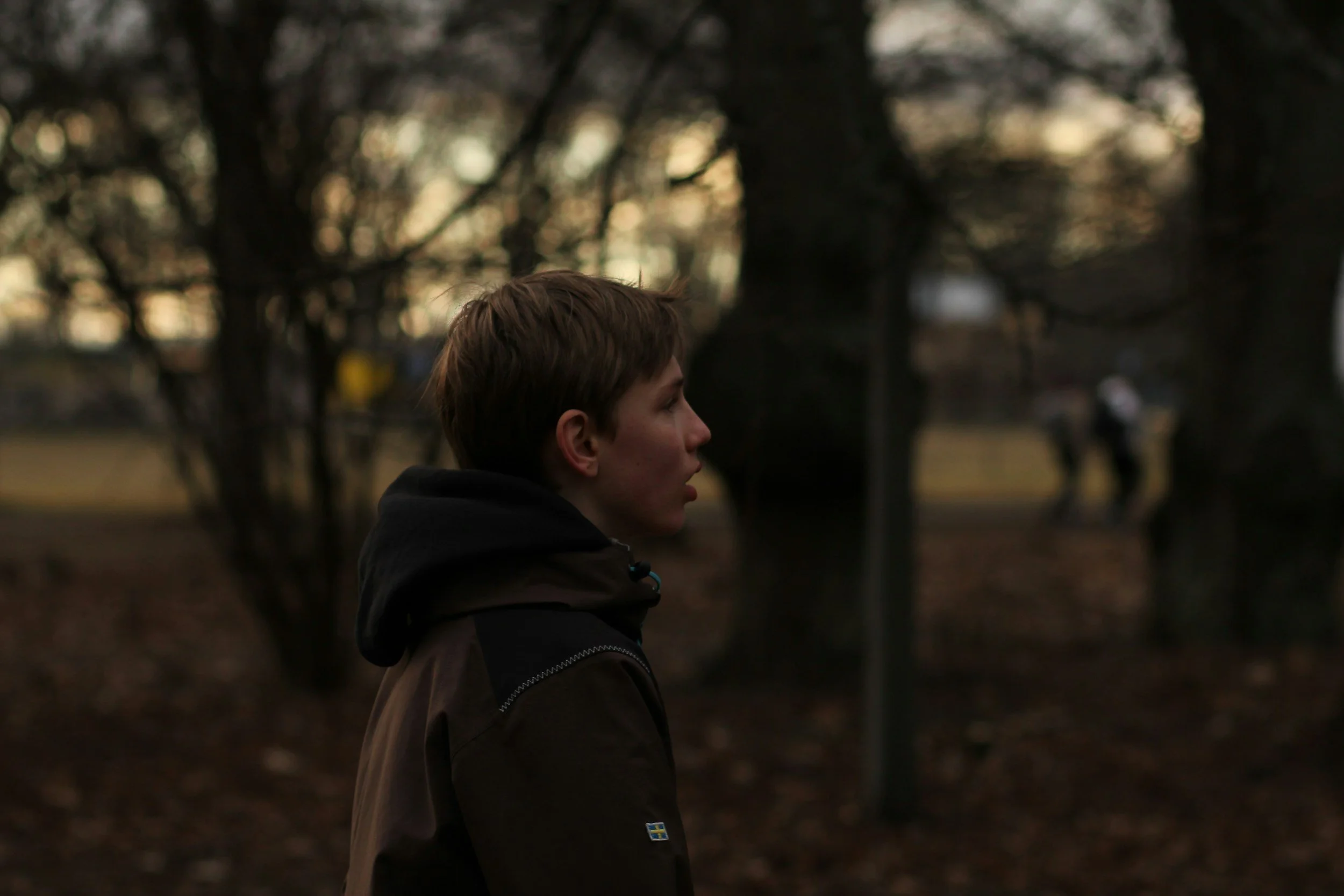 A young man standing outdoors in a park during dusk, with trees and blurred people in the background.