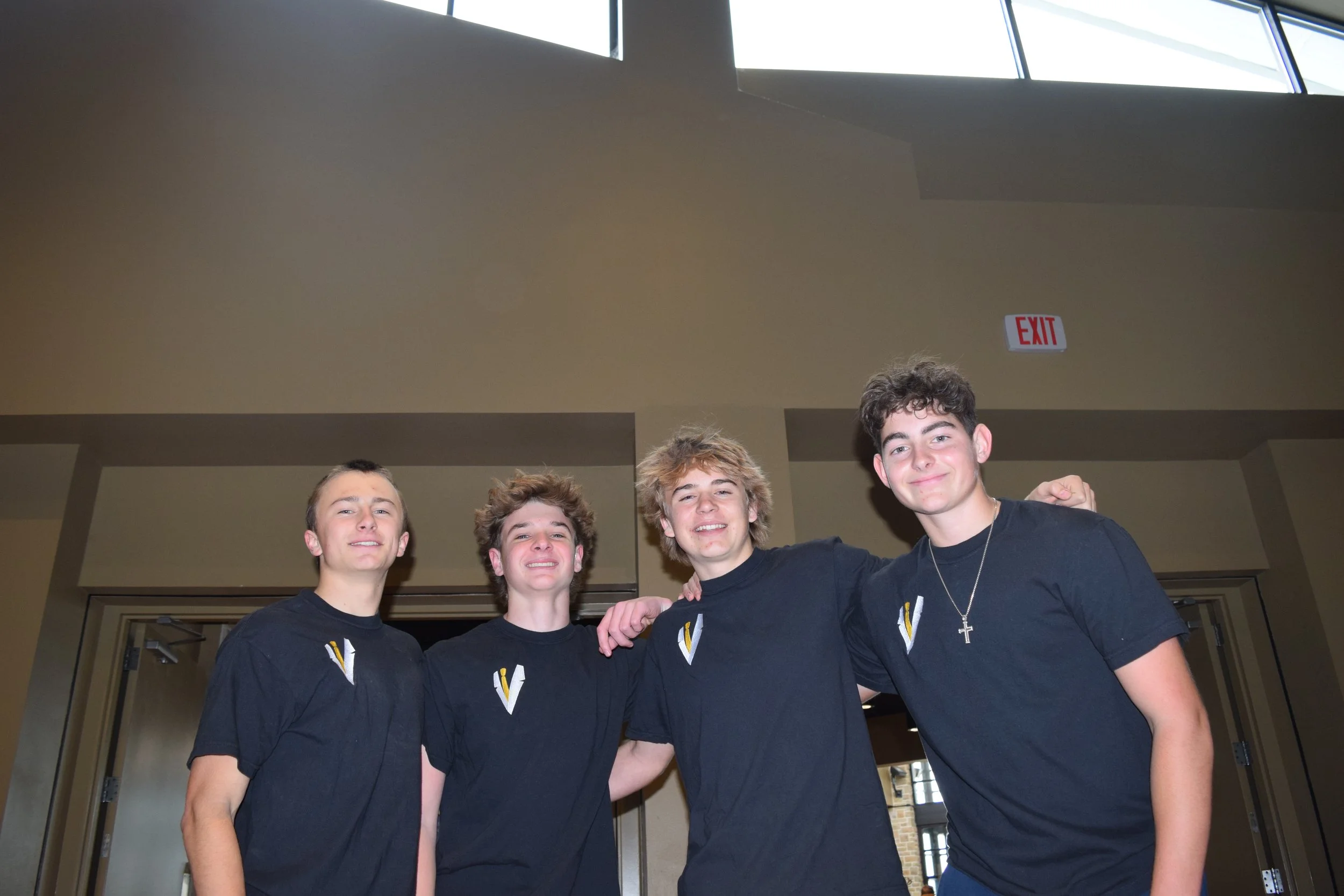 Four teenage boys standing together indoors, smiling, wearing matching black T-shirts with a logo on the chest, with their arms around each other's shoulders.