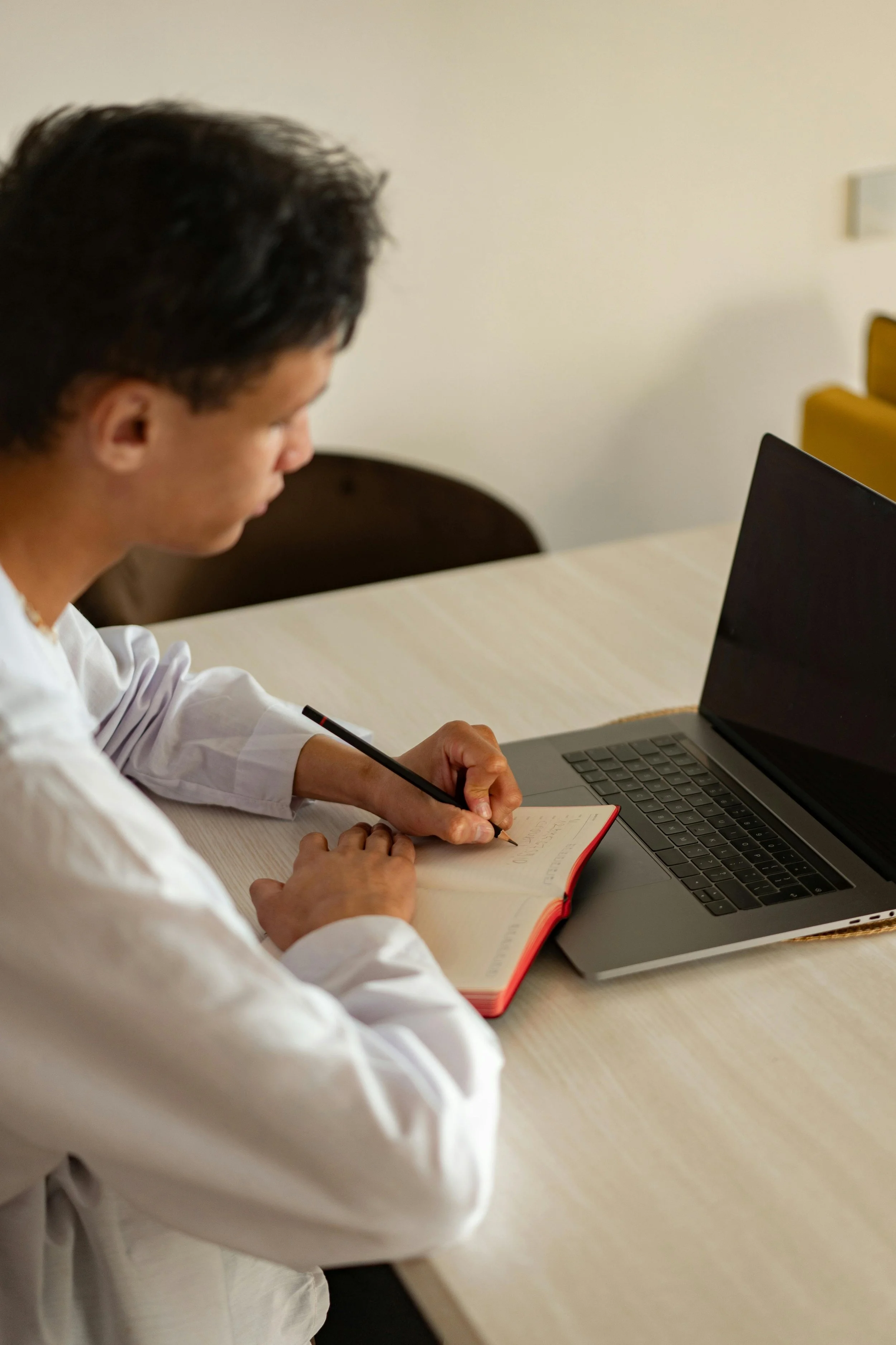 A person sitting at a table writing in a notebook with a laptop open beside them.