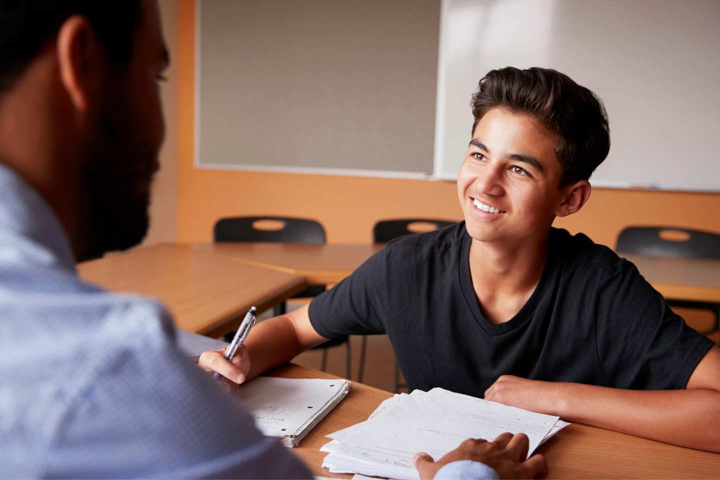 A young man with dark hair smiling during a conversation with an older man in a classroom or meeting room, with papers and a pen on the table.