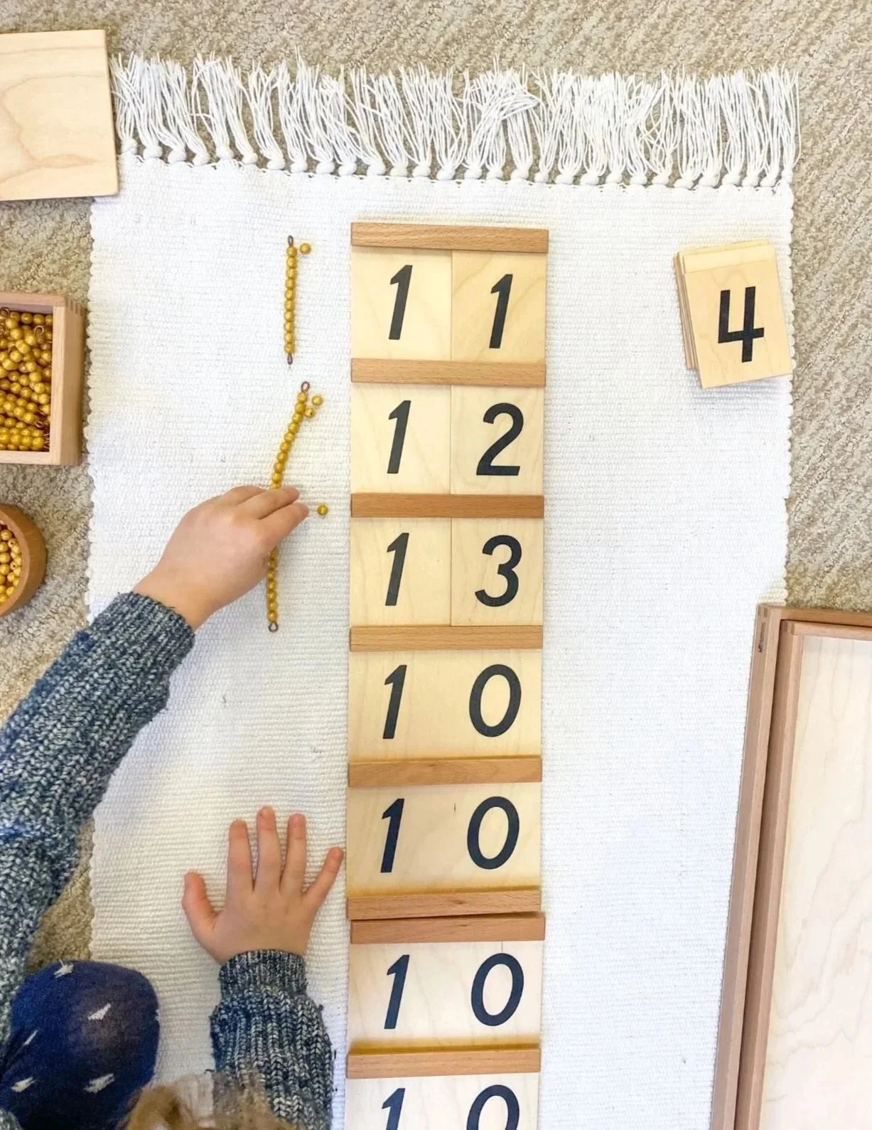 Child arranging yellow beads on a string next to wooden number blocks with black numerals from 11 to 10, set on a white rug, surrounded by small boxes with yellow beads, in a classroom or learning environment.