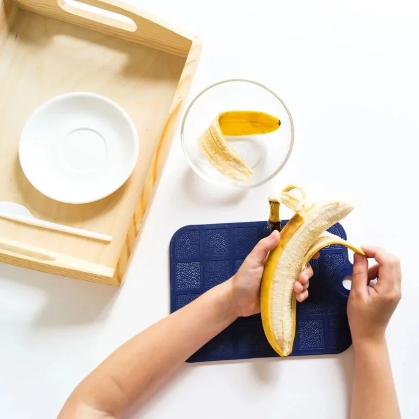 Person peeling a banana over a blue cutting board, with a glass of water and a wooden tray nearby.
